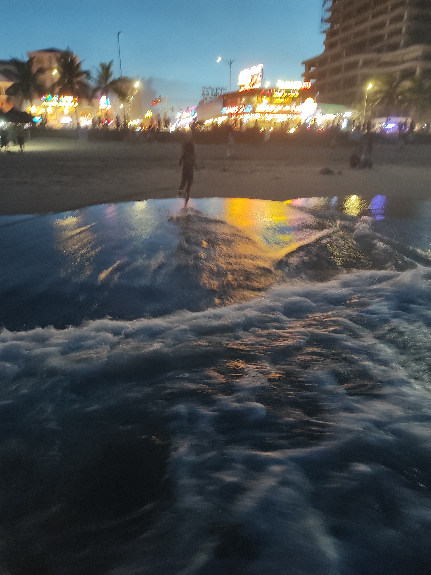 The image shows a beach in Đà Nẵng, Vietnam, at dusk. Waves are crashing in the foreground. In the midground, there is a person walking in the shallow water near the shoreline. In the background, there is a brightly lit cityscape with buildings, trees, and many people. A crescent moon is visible in the upper-left.