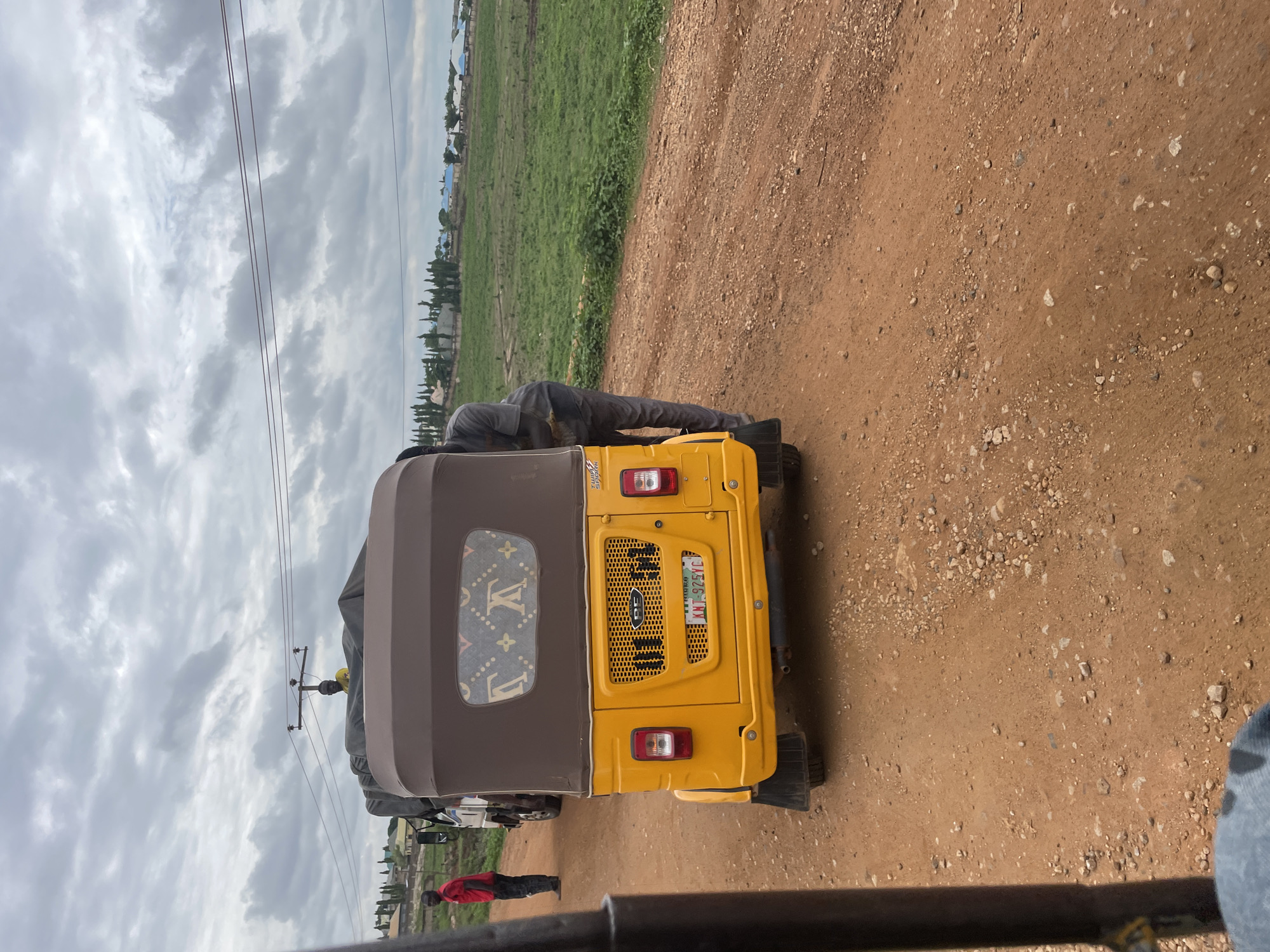In a rural setting in Unknown, Nigeria, a yellow tricycle with a brown canopy is parked on a dirt road. The tricycle has a license plate reading "KNT 925YC". Two men are present: one in a red shirt stands on the left side of the road, and another in a grey shirt is near the back of the tricycle. In the background, a green field stretches towards a cluster of houses with blue roofs, and a cloudy sky hangs overhead. Power lines are visible in the distance.