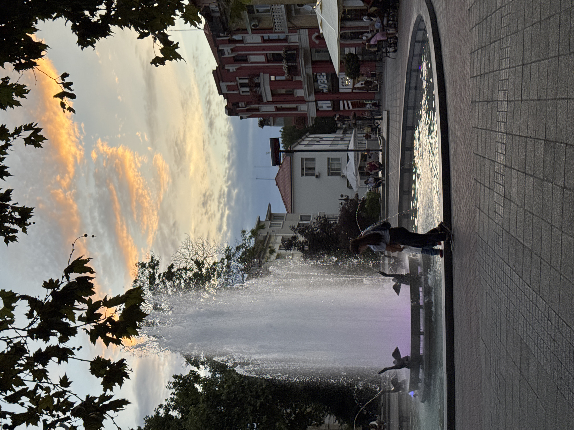 The image shows a wide fountain in a paved public square, with two people walking nearby. The fountain is large and actively spraying water, with decorative bird sculptures at its base. The water is illuminated with purple and pink lights. Behind the fountain, there are buildings in the background, one of which is a red building with white trim. The sky above is cloudy with streaks of orange and yellow, suggesting it is either sunrise or sunset. Trees with dark green leaves frame the top of the image. The paving stones in the foreground are a mix of grey and darker grey. The scene appears to be a lively urban public space. This fountain is located in the pedestrian zone of the city of Plovdiv, Bulgaria, specifically in the area of the main pedestrian street, Knyaz Alexander I.