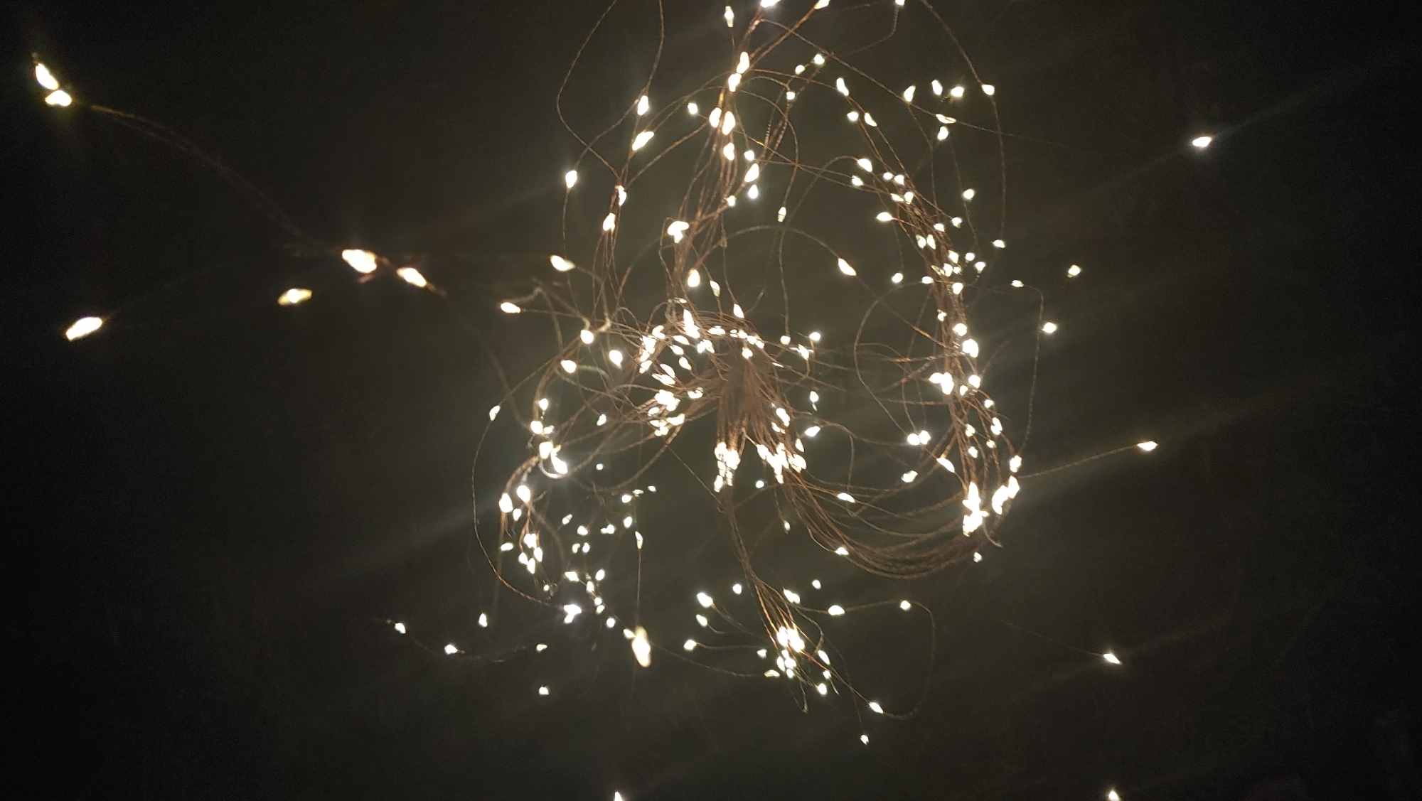The image shows a close-up of a string of fairy lights against a dark background, with some of the lights appearing as small, bright orbs and others as streaks due to motion blur or lens flare. The lights are small and white, attached to thin, wire-like branches that are arranged in a somewhat chaotic, wispy manner. The overall impression is of a decorative light installation, possibly outdoors at night. There is no discernible information in the image that would allow for the identification of the city of Mukachevo, Ukraine, or any specific location within it. The image is devoid of people or any context that would suggest a particular setting beyond a dark environment where lights are used for illumination or decoration.