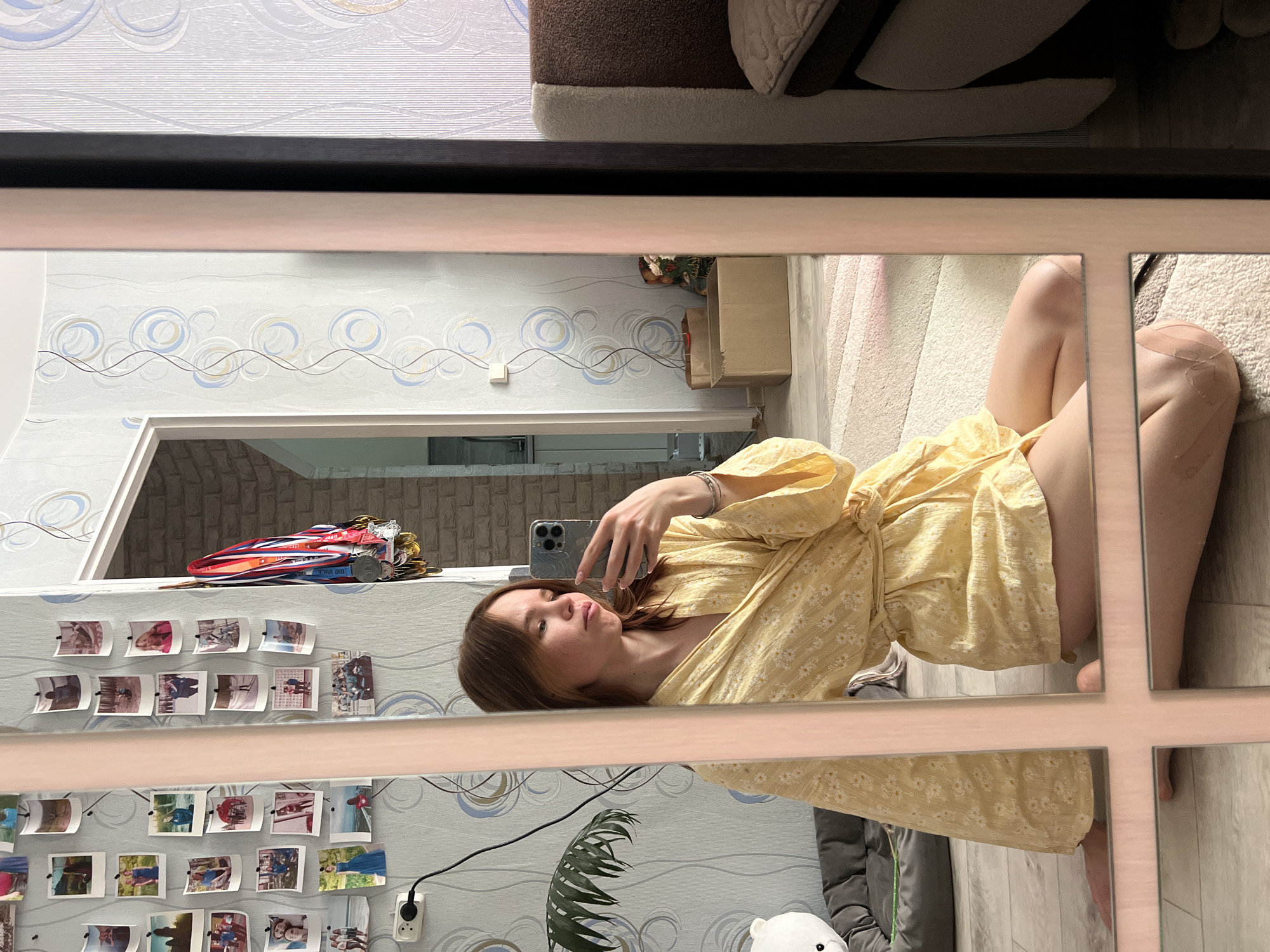 Here's a detailed description of the image:
**Scene:**
The image captures a reflection in a paneled mirror. In the reflection, a woman is visible. She is wearing a yellow floral dress and appears to be taking a picture of herself with a smartphone. She is seated on the floor.
**Surroundings:**
The room reflected in the mirror features patterned wallpaper with blue and gray swirls on a white background. A collection of photographs is displayed on the wall, attached with tape. Above the photos is a shelf holding medals and awards. An electrical outlet is visible near the bottom of the frame, along with a potted plant. A plush toy is also visible on the floor.
**Location:**
While the image provides interior details, there is insufficient visual information to confirm the location is in Chaikovsky, Russia. This conclusion cannot be definitively drawn from the image alone.
**People:**
The primary subject is a young woman holding a smartphone and posing for a photo. No other people are directly visible.
**Objects:**
The key objects include a smartphone, a yellow floral dress, a mirror, photos, medals, an electrical outlet, a potted plant, and a plush toy.
**Actions:**
The woman is taking a selfie using her smartphone. The other objects remain static within the scene.