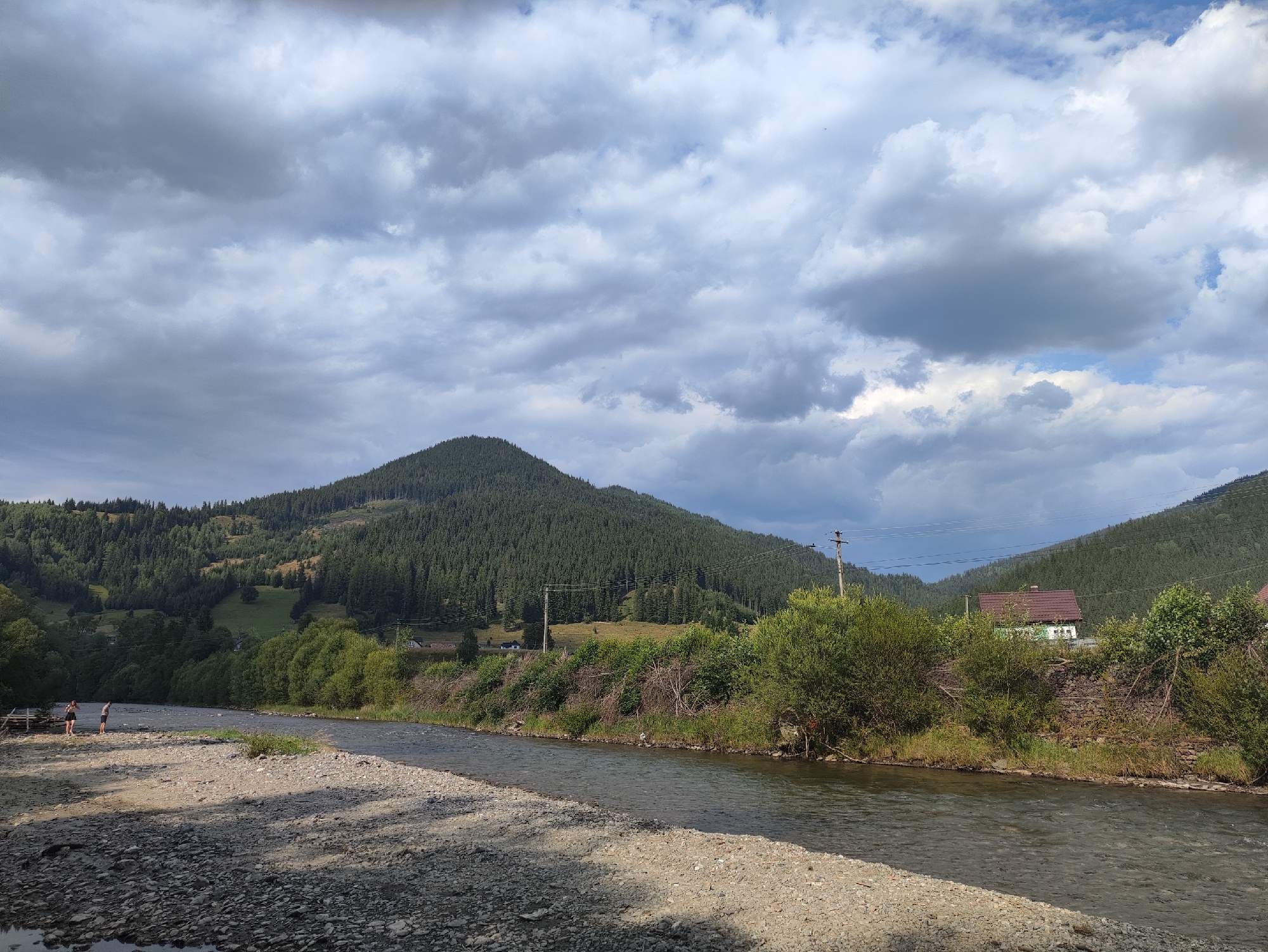 The image depicts a scenic view of a river flowing through a mountainous, forested landscape. In the foreground, a gravelly riverbank is visible, with two individuals standing near the water's edge. The river itself appears clear and shallow, with a rocky bed. Across the river, a lush green hillside is covered with dense evergreen trees, leading up to a mountain peak. In the middle ground, a small cluster of houses is nestled among the trees on the hillside. Power lines stretch across the scene, supported by utility poles. The sky above is a mix of heavy, gray clouds and patches of blue, suggesting a dynamic weather system. The overall impression is of a tranquil, natural setting, possibly a rural area or a park. The location is identified as being in the city of Botoș, Romania, though specific landmarks are not identifiable without further context.