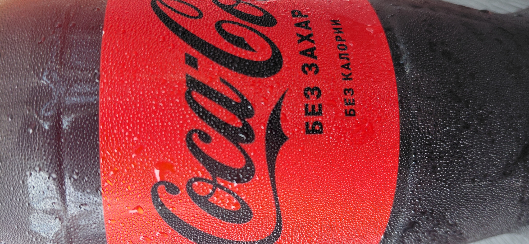 The image shows a close-up of a Coca-Cola bottle with condensation on its surface. The bottle has a red label with the Coca-Cola logo in black. Below the logo, there are two lines of text in black Cyrillic letters: "БЕЗ ЗАХАР" (without sugar) and "БЕЗ КАЛОРИИ" (without calories). The background is dark and textured, also showing condensation. The image does not contain any information or context related to the city of Bozhurishte, Bulgaria.