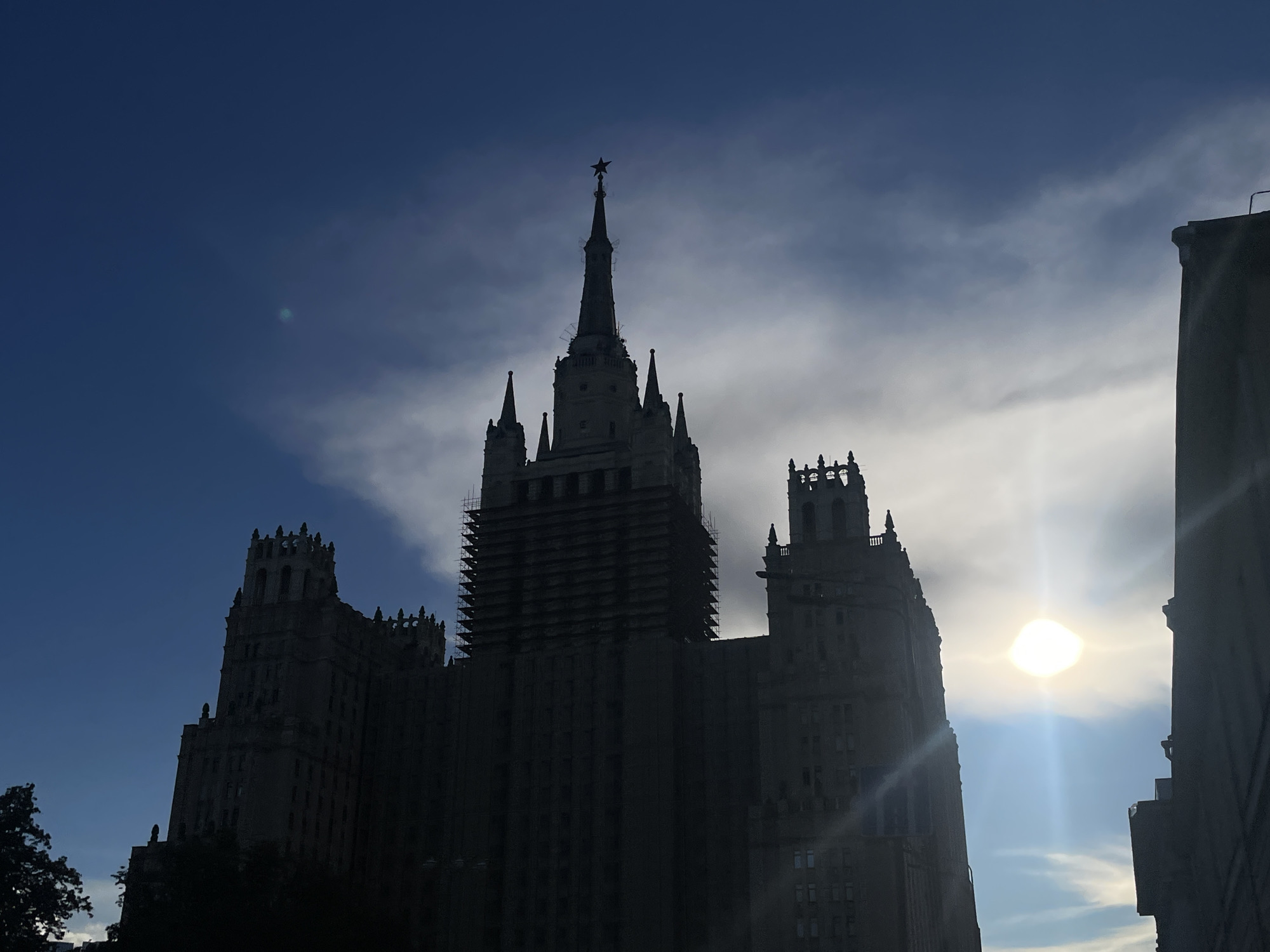 The image depicts the Kotelnicheskaya Embankment Building in Moscow, Russia, silhouetted against a bright sky with scattered clouds. The sun is visible as a bright, blown-out orb in the upper right portion of the frame. The iconic Stalinist skyscraper, a prominent landmark in the city, dominates the center of the image. Its intricate architectural details, including spires and decorative elements, are visible in silhouette. The lower part of the building is partially obscured by dark trees. The overall scene suggests a late afternoon or early morning light condition, with the sun's rays creating a lens flare effect. The sky exhibits a gradient of blues and grays, with some wispy clouds suggesting movement or atmospheric conditions. No people are visible in the image. The perspective is from a low angle, looking up at the towering structure.