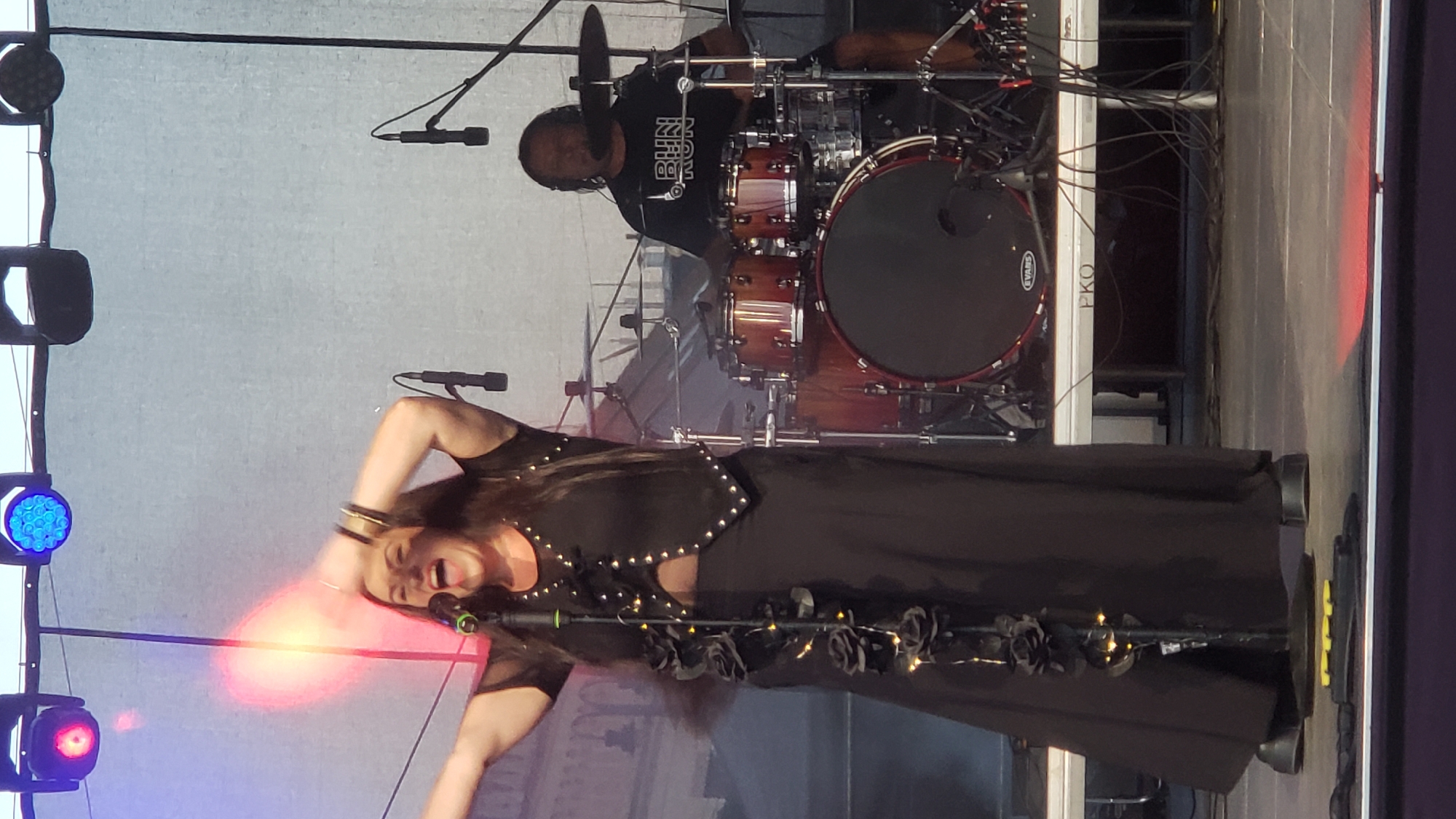 The image shows a female singer on a stage in front of a drum set. She is wearing a black, sheer top with silver studs and a long black skirt. She has dark hair and is singing into a microphone with her arms raised. The drum set is behind her, and a male drummer with headphones is playing. The stage has lighting rigs above, with red and blue lights visible. In the background, there is a large white tarp. The stage also appears to be set up outdoors, possibly during a festival or concert. The location is identified as Prešov, Slovakia.