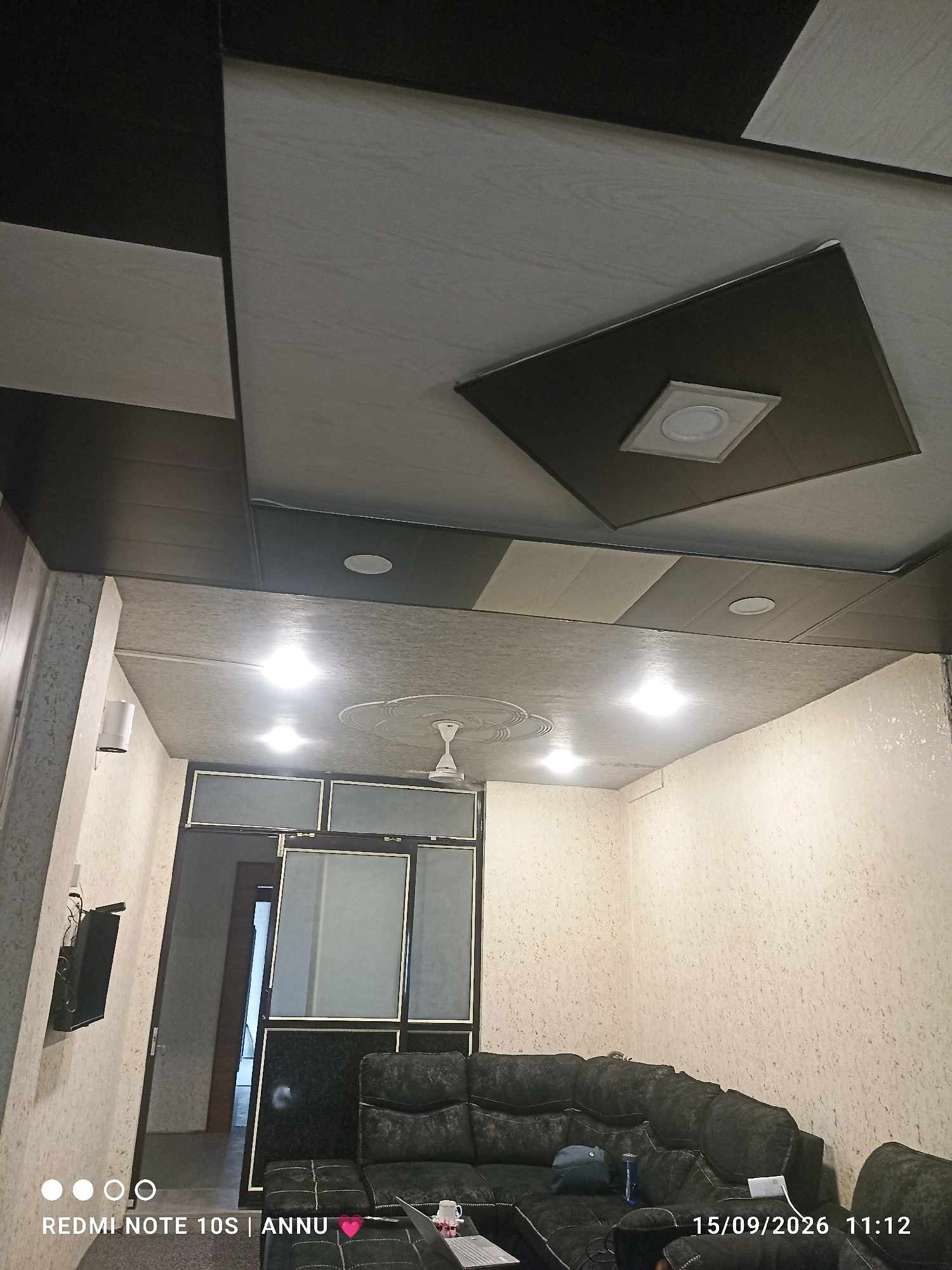 The image shows the interior of a room with a patterned ceiling and textured walls. The ceiling features a geometric design with alternating black and grey panels, some of which are recessed and contain light fixtures. One prominent recessed area has a square frame surrounding a round light. A ceiling fan is visible in the lower right portion of the image. The walls are covered in a light-colored textured wallpaper. In the foreground, a dark-colored sectional sofa is present, with a few items on it, including a blue backpack, a water bottle, and a white power bank. A laptop is visible on a low table in front of the sofa. To the left of the sofa, a wall-mounted television is visible. A doorway is visible in the background, leading to another part of the interior. The overall impression is of a modern living space. The image is dated 15/09/2026 and time-stamped 11:12. The text "REDMI NOTE 10S | ANNU" with a pink heart emoji is overlaid on the bottom left of the image. Based on the provided metadata and context, this image is from Pampore, India.