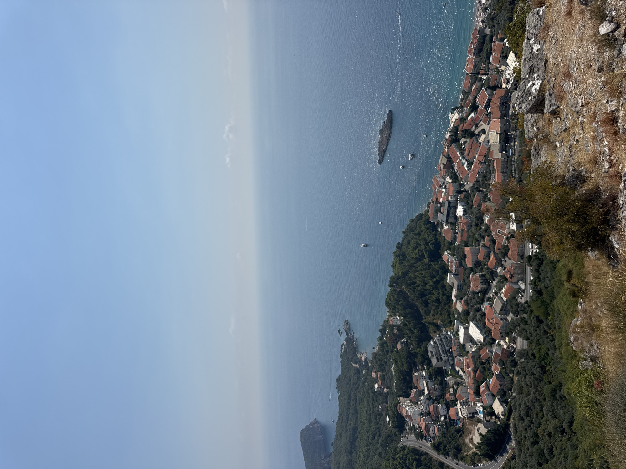This aerial view captures a section of Sveti Stefan, Montenegro, showcasing a coastal town nestled amidst lush green hills and overlooking the Adriatic Sea. The foreground is dominated by rocky terrain with sparse vegetation, hinting at a vantage point from a higher elevation. Below, a densely packed settlement with characteristic red-tiled roofs spreads along the coastline. Buildings of varying sizes and architectural styles are visible, interspersed with olive groves and trees. A winding road curves through the town, with a few cars apparent. The sea, a vibrant blue, stretches towards the horizon, dotted with a few boats, including a larger vessel anchored offshore and a small island or rock formation. The sky is a clear, pale blue with a few wispy clouds. The overall impression is one of a picturesque coastal community bathed in bright sunlight.