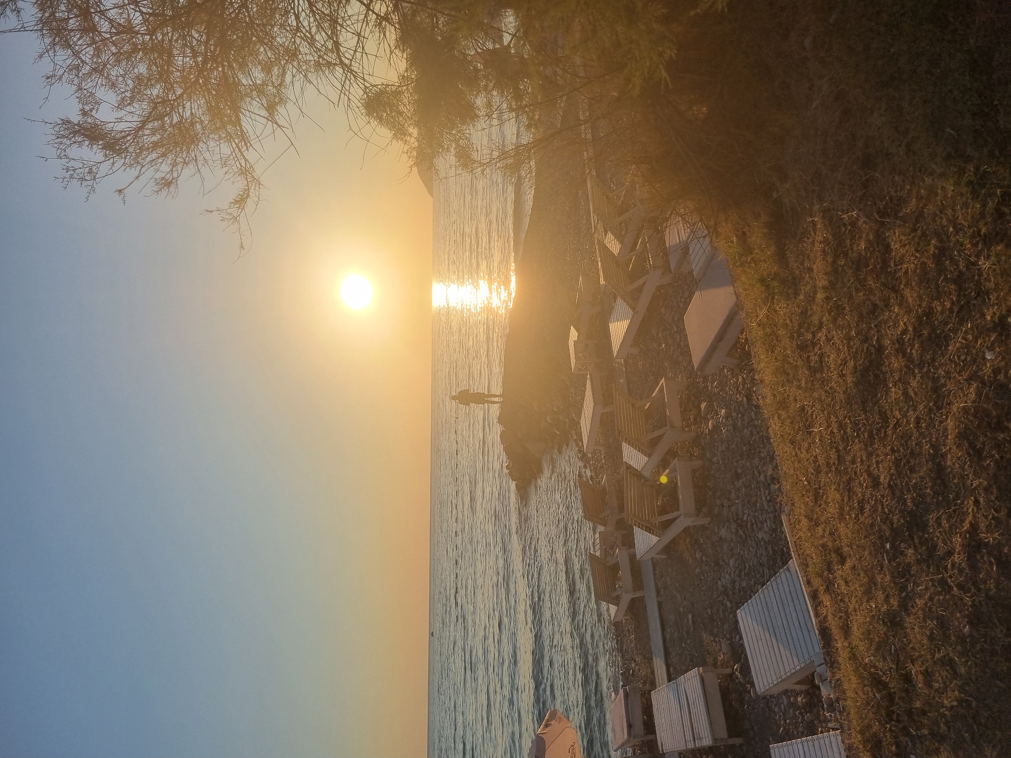 The image depicts a beach scene at sunset in Bar, Montenegro. The sun is low on the horizon, casting a warm glow and creating a reflection on the calm sea. A lone figure stands on a rocky outcrop at the edge of the water, silhouetted against the bright sky. Beach chairs are arranged on the pebbled shore, some in rows and others scattered. A tree with sparse branches is visible in the foreground on the right, its foliage backlit by the setting sun. The sky transitions from a pale blue at the top to a soft yellow and orange near the horizon. The water appears tranquil with gentle ripples. The overall atmosphere is serene and peaceful.