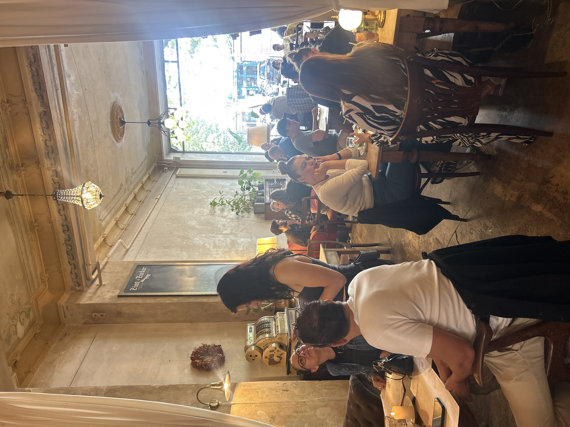 The image depicts the interior of a bustling cafe in Berlin, Germany, featuring a variety of patrons engaged in dining and conversation. The cafe's ambiance is characterized by its vintage decor, with exposed brick walls, ornate ceiling moldings, and a combination of rustic and classic furniture.
In the foreground, a group of people are seated around a table, engrossed in their meals and drinks. A woman in a white t-shirt is facing away from the camera, while a woman in a black and white patterned dress is seated in a wooden chair, her back also to the viewer. Another woman with dark hair, wearing a grey tank top, stands and interacts with a woman wearing glasses and a denim jacket. A vintage cash register is visible on a shelf behind them.
The midground reveals more diners scattered throughout the cafe, some standing and others seated. Natural light streams in from a large window at the back, illuminating the space and offering a glimpse of the street outside, where cars and pedestrians are visible.
The background showcases the cafe's architectural details, including chandeliers hanging from the ceiling and a menu board mounted on the wall. The overall scene conveys a lively and social atmosphere, typical of a popular urban cafe.