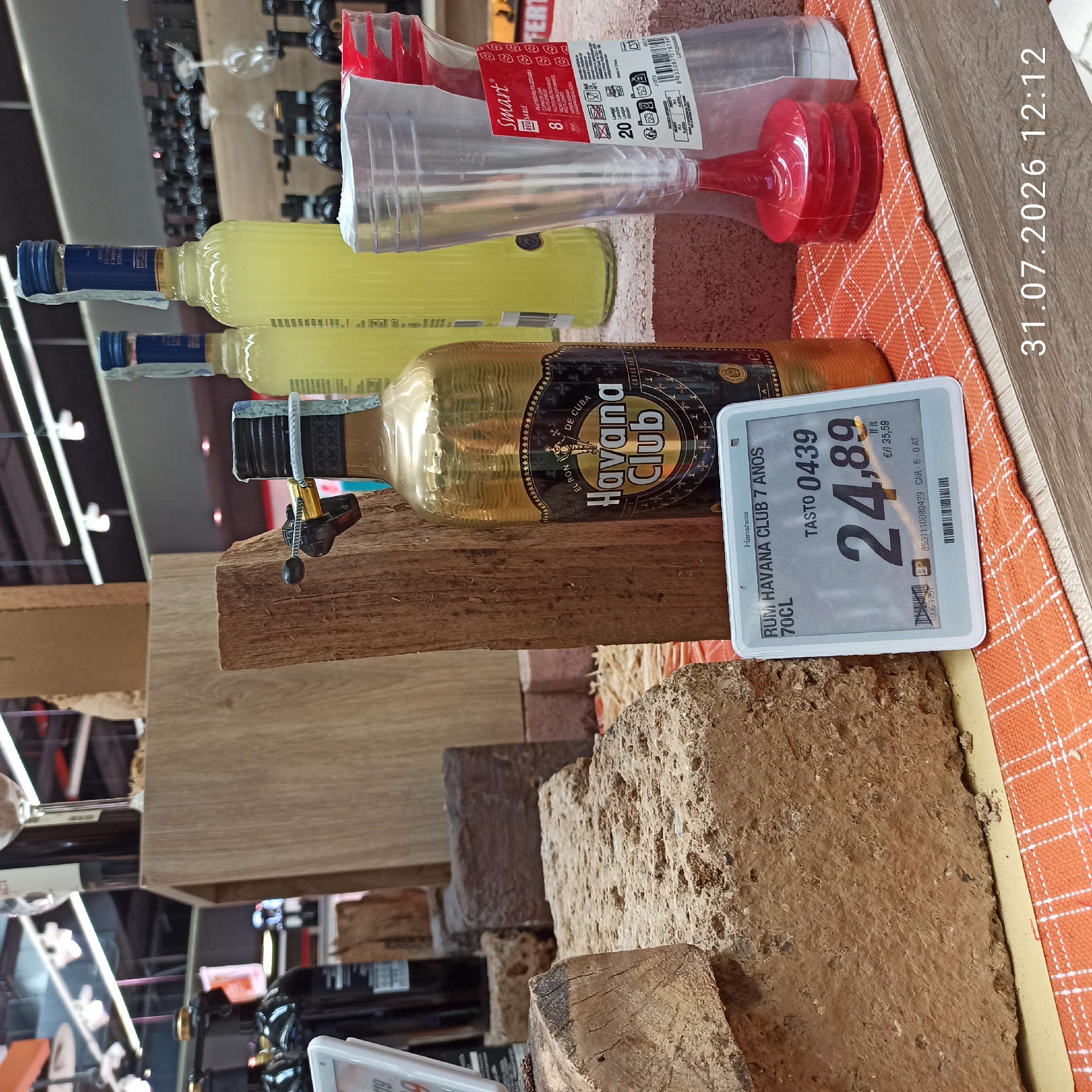 The image depicts a retail display of alcoholic beverages, specifically rum and what appears to be a liqueur, in a store located in Modugno, Italy. The foreground features a price tag displaying "RUM HAVANA CLUB 7 ANOS 70CL" with a price of "24,89 €/lt 35,59". Next to it is a partially obscured bottle of Havana Club rum, its gold foil label prominently visible. To the right, a package of disposable plastic flutes is displayed. In the background, several bottles of yellow liquid, likely liqueur, are arranged on a shelf, along with other dark-colored bottles. The display is set against a backdrop of wooden planks and textured surfaces, suggesting a rustic or artisanal theme. The overall impression is of a point-of-sale display within a supermarket or liquor store. The timestamp in the bottom right corner indicates the image was captured on July 31, 2026, at 12:12.