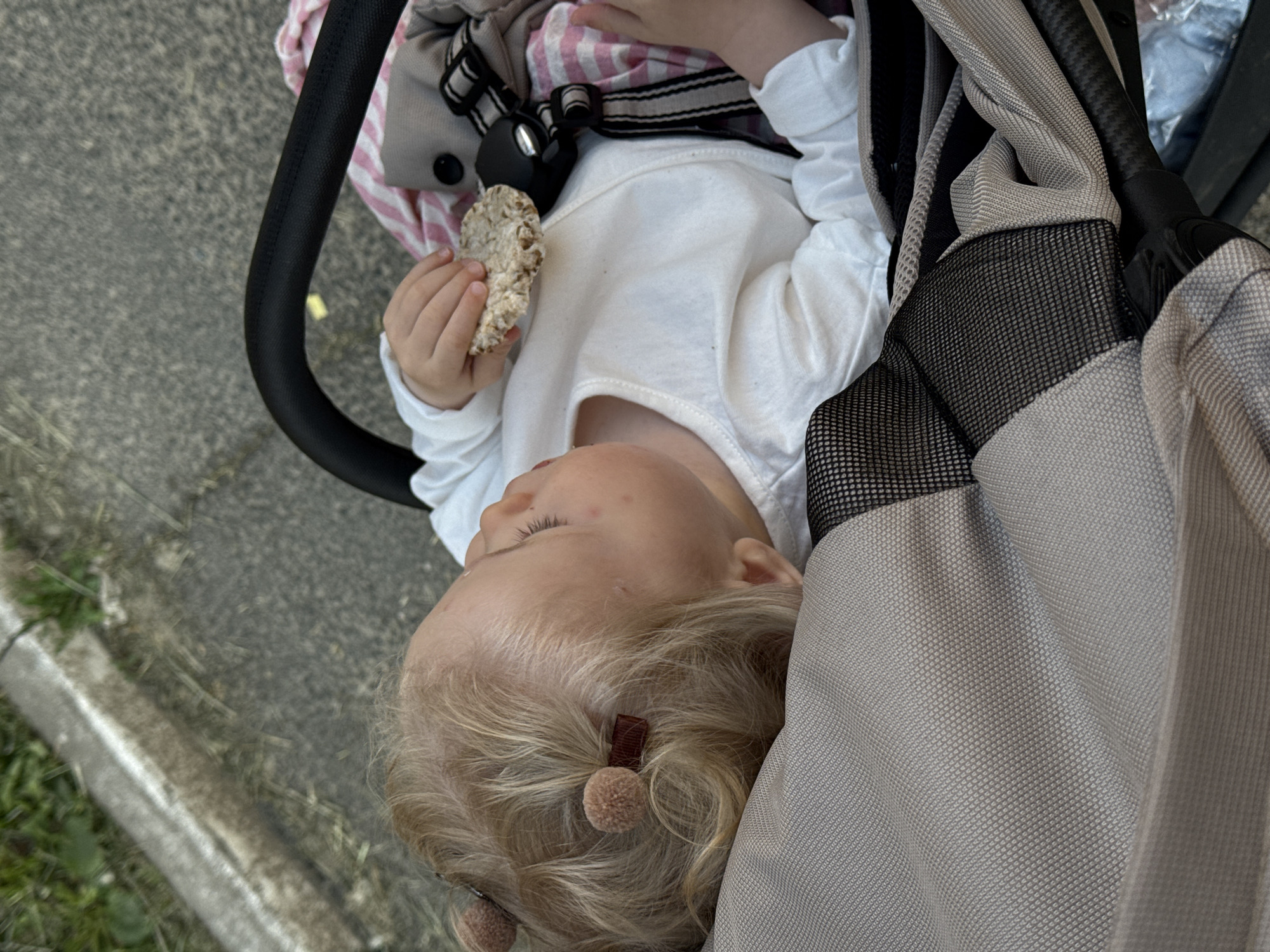 A fair-skinned toddler is lying in a stroller while holding a rice cake in their left hand. The child has blonde hair styled with two small brown hair ties with pom-poms. They are wearing a white long-sleeved shirt. The stroller has a black handle and a grey and black canopy. The stroller is on pavement. The location is in the city of Vladimir, Russia.