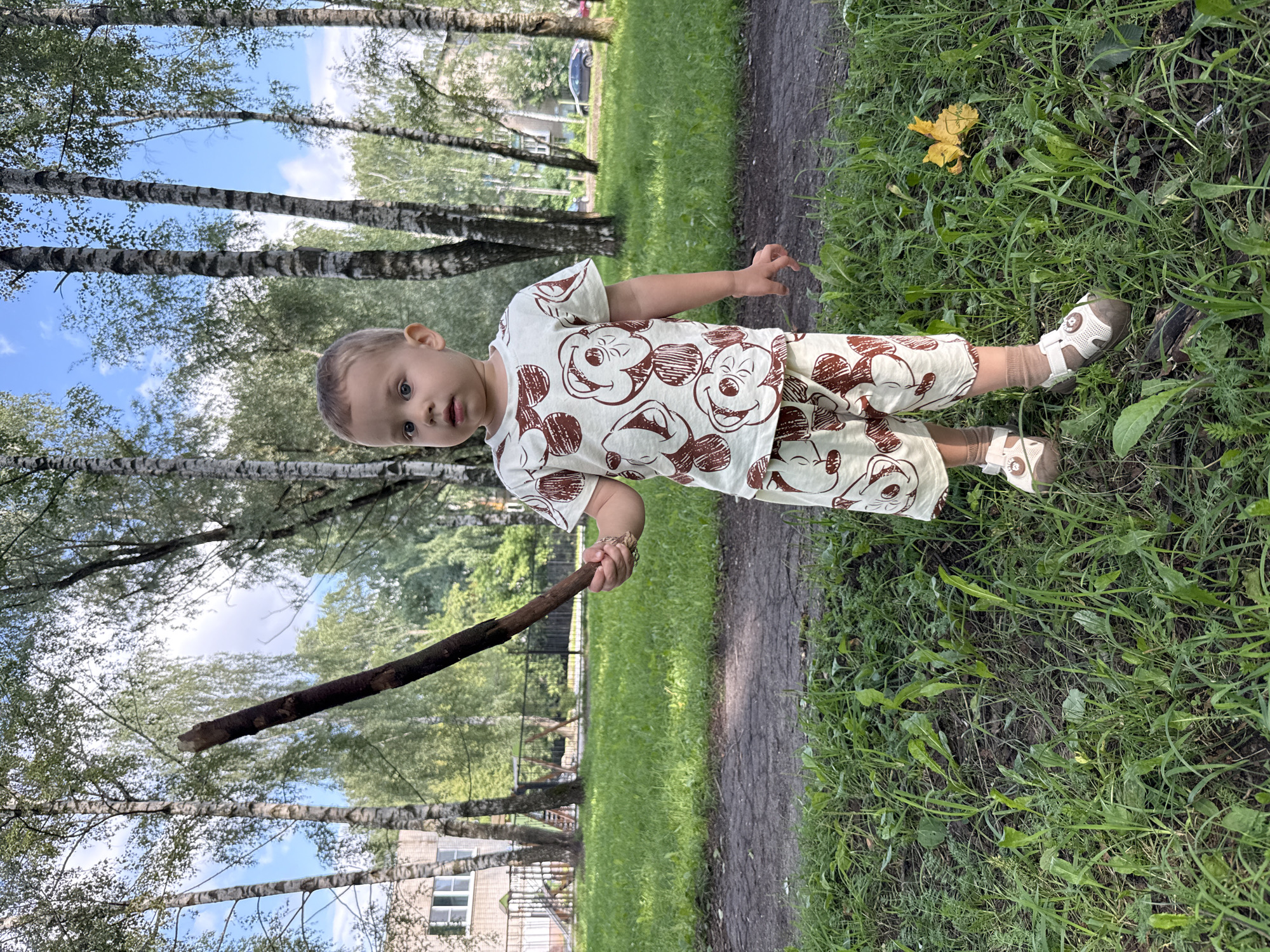 A young boy stands in a grassy area outdoors, holding a dark brown stick. He is wearing a light-colored t-shirt and shorts with a repeating pattern of brown circular shapes, possibly flowers or stylized faces. He also has on light-colored sandals with brown accents and socks. The background features birch trees with striped bark and lush green foliage. In the distance, a building with windows and a fence can be seen, along with a parked car. The ground is covered with grass and a dirt path. The sky is blue with scattered white clouds. The image appears to be taken in the city of Rusanovo, Russia, based on the visual cues and context provided.