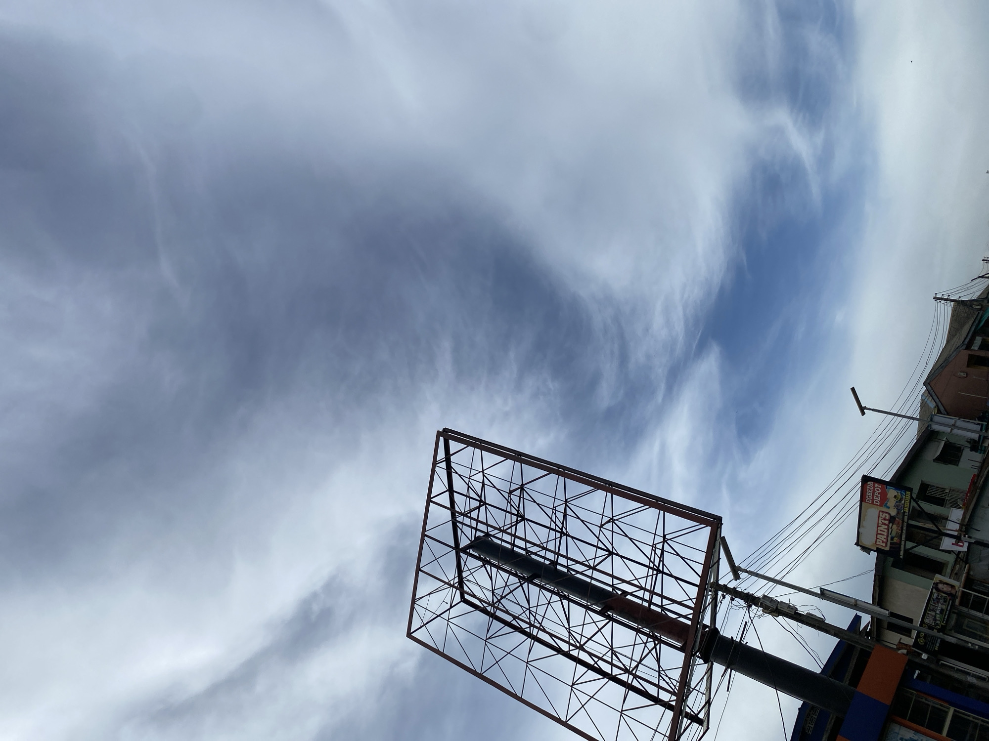 The image captures an upward-looking view of an urban sky in Lagos, Nigeria, dominated by a large, empty billboard frame in the foreground. The sky features a mix of wispy white and grey clouds against patches of light blue, suggesting a partially cloudy daytime.
The main subject is a large, rectangular billboard frame made of dark metal scaffolding, angled slightly, with its base extending out of the frame towards the lower right. It appears to be an advertising structure awaiting a banner. To the right of this frame, a utility pole stands tall, strung with numerous electrical wires that stretch across the mid-ground. Attached to the pole are various components, including what appears to be a street light fixture.
Further to the right, parts of urban buildings are visible. One building, possibly painted light green and off-white, features a smaller billboard attached to its side. This billboard clearly displays the text "EGBEDA DEPOT" at the top, with the word "PAINTS" prominently beneath it. Smaller, less legible text below "PAINTS" seems to mention "HOUSEHOLD," "INDUSTRIAL," and "AUTOMOTIVE" paint types, followed by "DELIVERY AVAILABLE." The overall scene suggests a bustling commercial area, typical of urban Lagos, with visible infrastructure and advertising. No people are visible in the frame, and the activity is limited to the static depiction of the urban environment under an open sky.