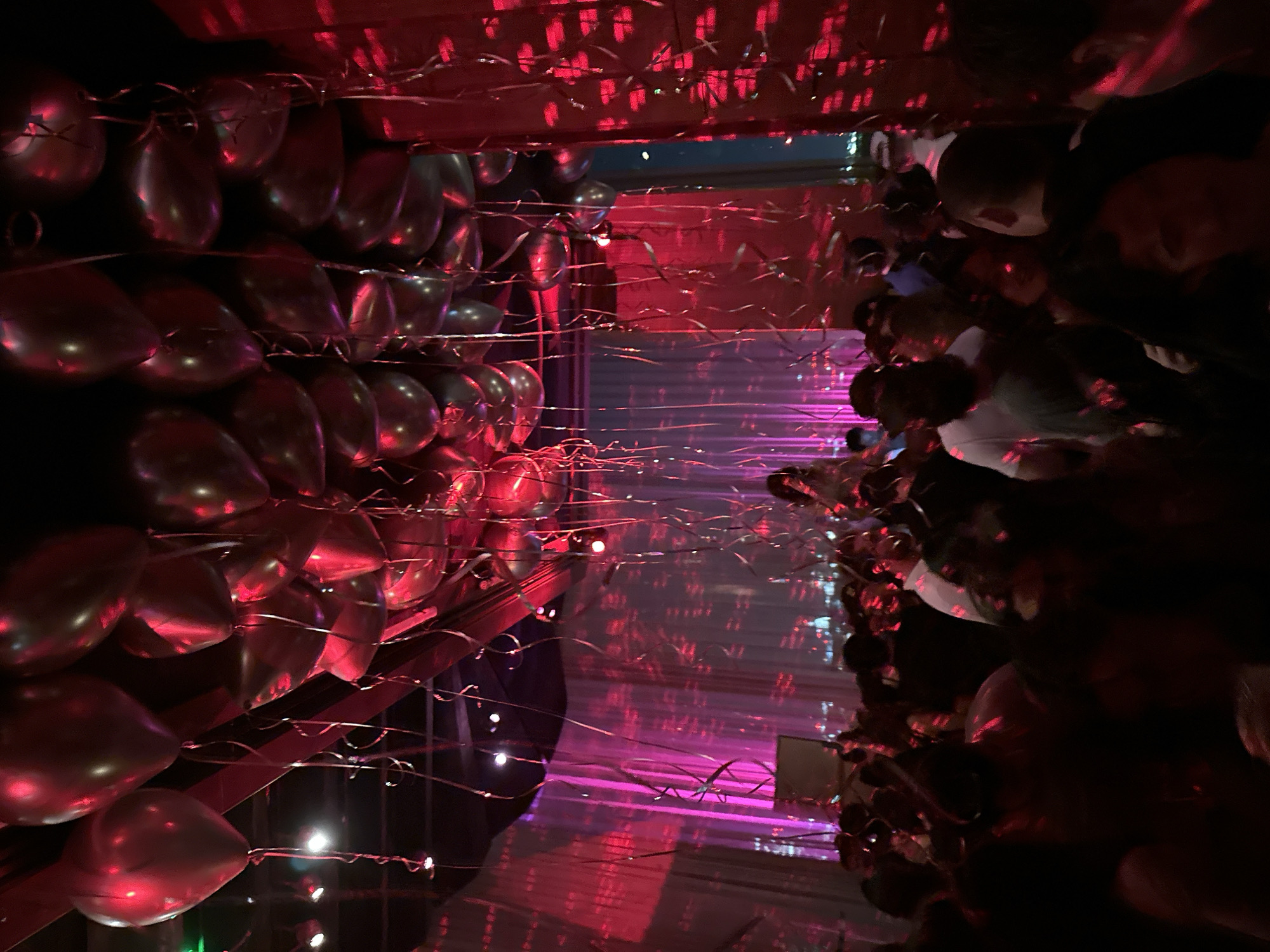 The image shows an indoor scene with dark, moody lighting primarily in shades of red and purple. There appear to be metallic balloons strung along the left side of the frame, possibly forming a decorative wall or backdrop. Thin, shimmering strands, possibly ribbon or tinsel, are draped across the scene. On the right side of the image, indistinct figures of people are visible. The overall ambiance suggests a party or event setting. Based on the red lighting and distinctive balloon arrangements, it is highly probable the location is a venue in London, United Kingdom, but definitive confirmation without additional visual cues is not feasible.