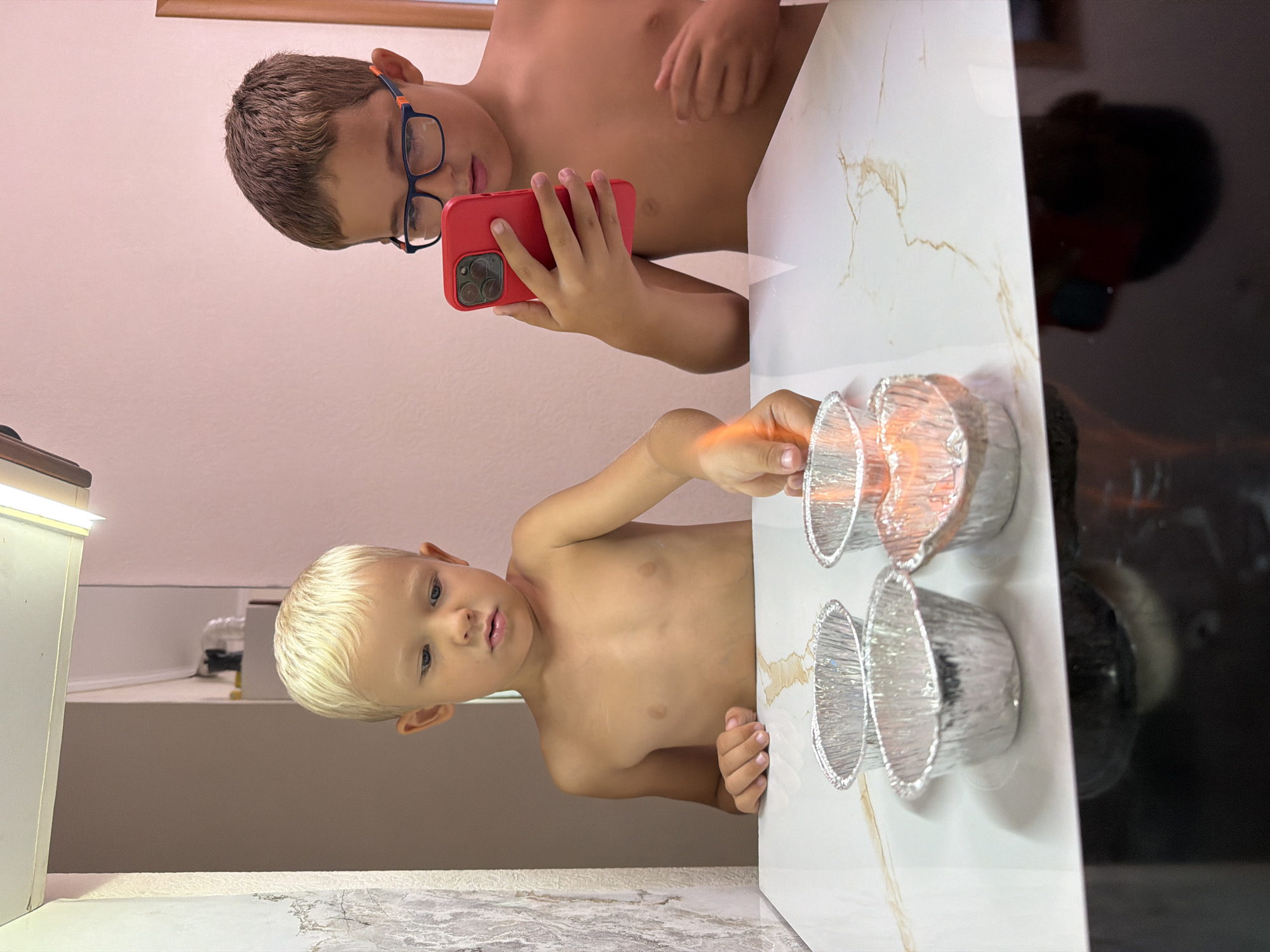 In a bright, indoor setting, two young boys are engaged in an activity. The older boy, wearing glasses and a red phone case, is holding a smartphone. The younger boy, with blonde hair, is reaching out towards several small, foil baking cups. One of these cups appears to have a lit candle inside, casting a warm glow. The scene is captured from a low angle, with the subjects appearing in the foreground. The background includes a light-colored wall and a dark, reflective surface, possibly a countertop or appliance. The overall impression is one of a casual, playful moment between the two children. The lighting suggests it is daytime. There is no visible text in the image.