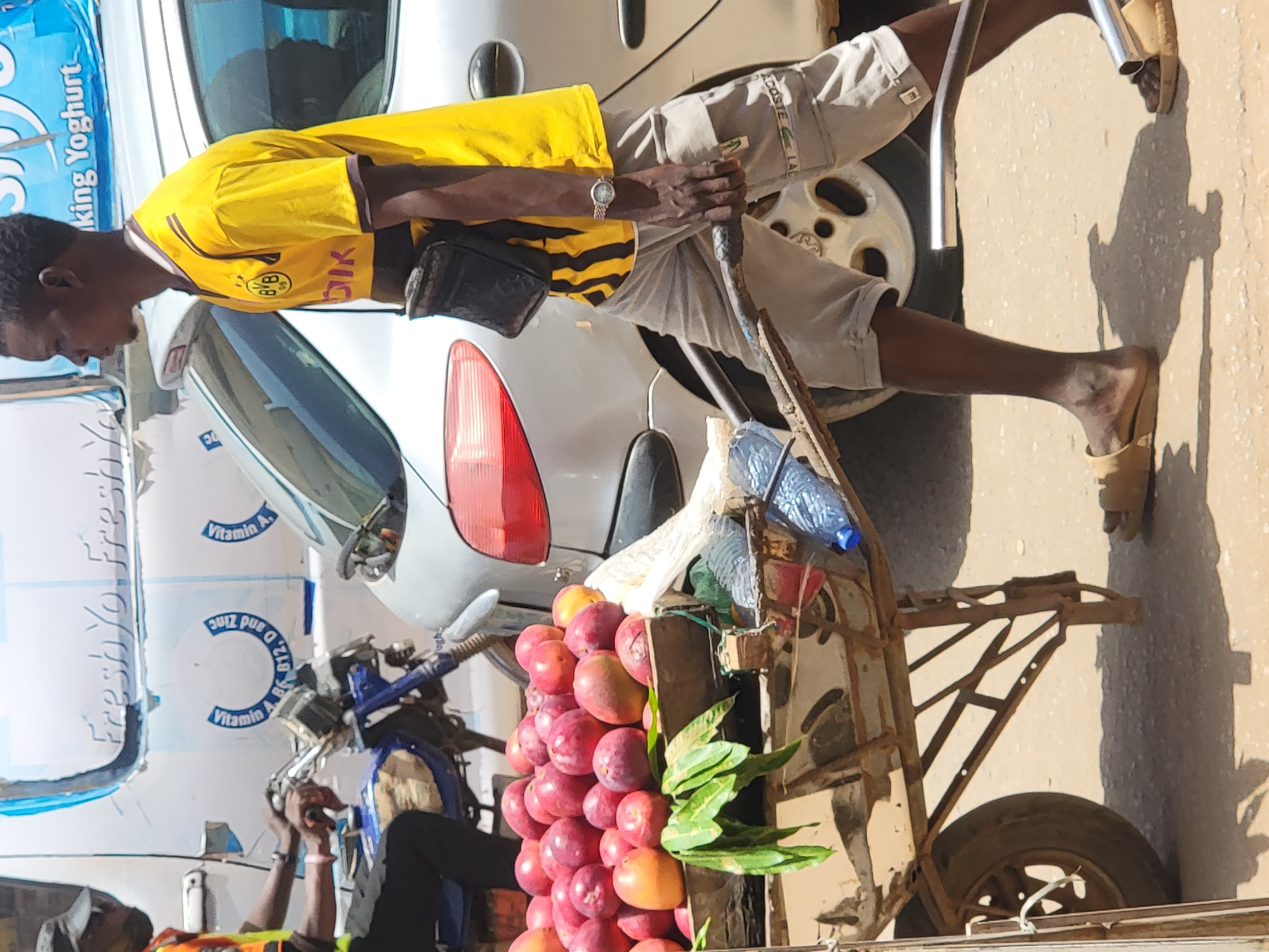This vivid outdoor scene, captured in bright daylight, depicts a bustling street, likely in Maiburiji, Nigeria. The image is centered on a man, presumably a street vendor, standing beside a mobile fruit cart. He wears a yellow and black striped short-sleeved jersey, prominently featuring the "BVB 09" logo and a partial "Dik" (likely from Borussia Dortmund), light-colored shorts with a "LACOSTE LAC" logo, and light-brown sandals. A wristwatch is visible on his left wrist as he holds onto the cart's frame.
The rustic metal and wooden cart is laden with a large pile of red-orange fruits, resembling plums or similar stone fruits, interspersed with some green leaves. A plastic water bottle rests on the cart's upper structure.
In the background, a silver car is partially visible, and behind it, a large white delivery vehicle (possibly a yogurt truck) displays blue branding. Visible text on this vehicle includes "Freshyo Fresh Ya king Yoghurt" and mentions "Vitamin A. B6 B12. D and Zinc." Below the car, parts of a blue motorcycle or scooter are visible. Another person, wearing a cap and an orange vest, is partially seen in the lower-left corner, seemingly engaged in an activity or resting. Strong, well-defined shadows cast on the light-colored ground indicate a sunny day, characteristic of the provided location context. The scene captures everyday commerce in an urban or semi-urban setting.