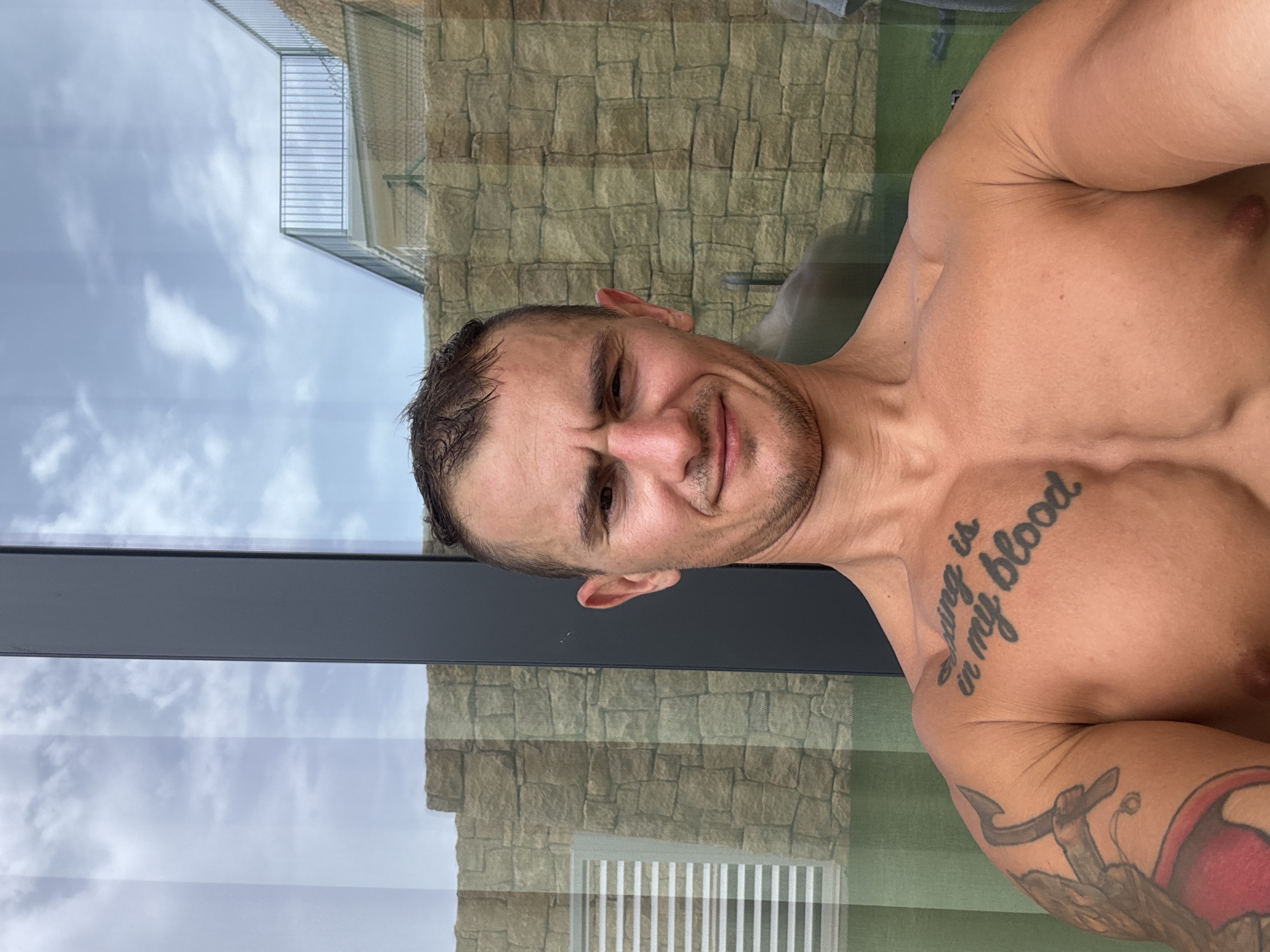 A man with short brown hair and a muscular build is taking a selfie. He is shirtless, revealing a tattoo on his chest that says "Living is in my blood" and another tattoo on his left arm. He has a faint smile and appears to be outdoors, with a stone wall and a balcony visible in the background. The location appears to be Finestrat, Spain, possibly a hotel or residential property. The lighting suggests it is daytime.