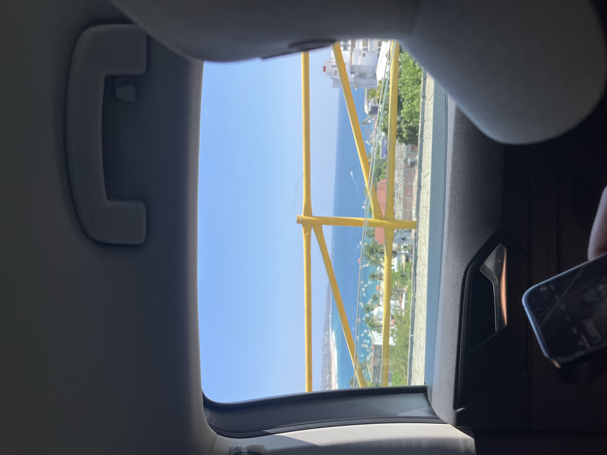 The image depicts a view from inside a car, looking out through an open sunroof. The location is the city of Puerto Escondido, Mexico. The scene visible through the sunroof features a clear blue sky, the ocean, a beach, and a town on a hill. There is a yellow scaffold on a building. Parts of the car's interior are visible, including a passenger handle and a section of the dashboard and door. The corner of a phone is visible in the lower right corner.