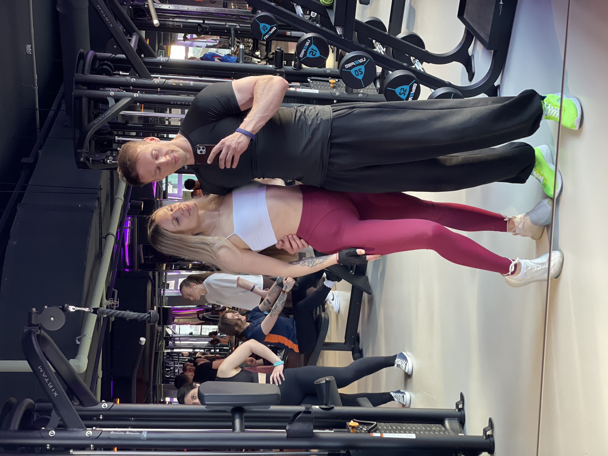 A full shot, taken at eye-level with a smartphone, shows a couple taking a mirror selfie in a gym. The couple stands in the foreground, looking at their phones. The male subject is on the right, wearing a black t-shirt and black sweatpants. He is holding his phone up to take the photo, with his left arm around the female subject. The female subject is on the left, wearing a white sports bra and pink leggings. She is looking at her phone and smiling.
In the background, several other gym-goers are visible, working out on various machines. The gym is well-lit and modern, with a variety of weight machines and free weights. The floor is a light-colored, polished surface, and the ceiling is dark with exposed pipes and lighting fixtures. Purple accent lighting is visible along the ceiling. The overall impression is that of a busy, upscale gym.