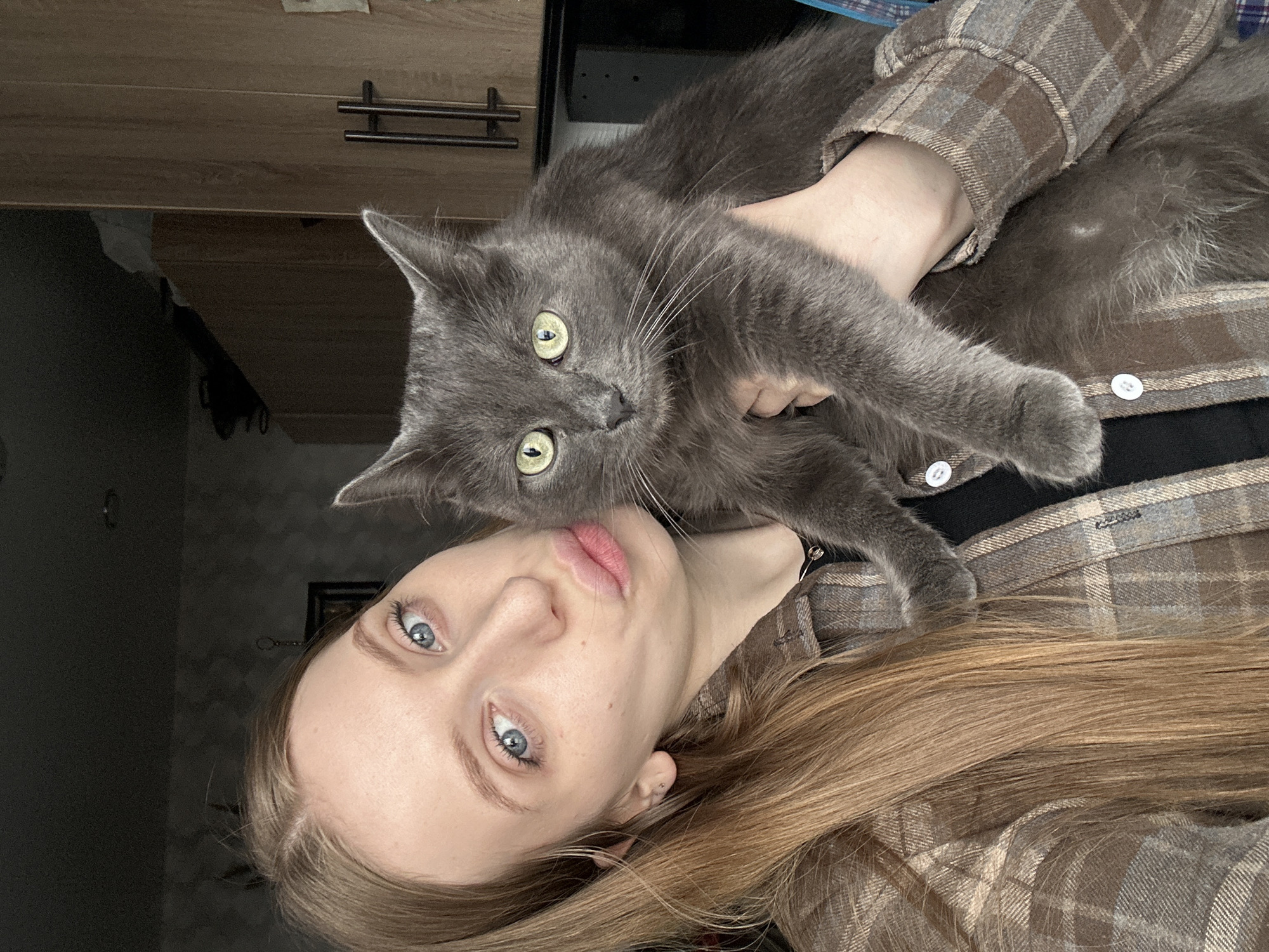 A young, fair-skinned woman with light blue eyes and long, light brown hair is holding a grey cat. The cat has yellow eyes and is looking at the camera. The woman is wearing a brown and grey plaid shirt over a black garment. The background includes a light brown cabinet with dark handles. The location is not possible to determine.