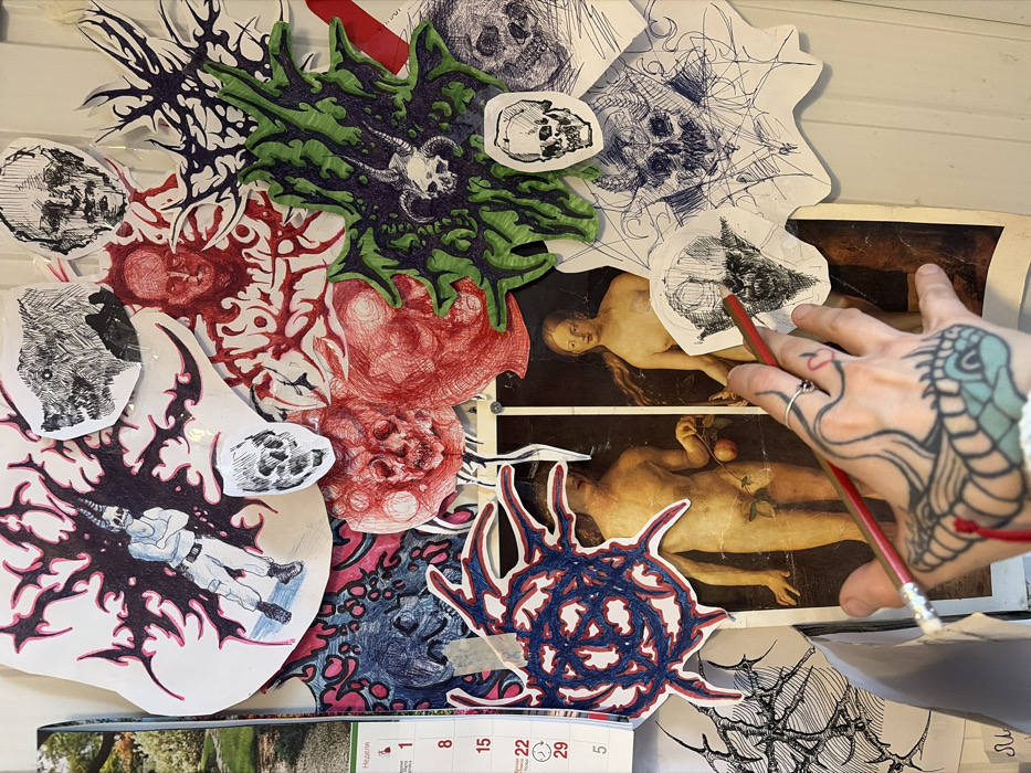 The image depicts a densely covered vertical surface, likely a wall or display board, located in Sofia, Bulgaria. The surface is adorned with a collage of cut-out drawings, prints, and a calendar segment.
Centrally, a faded print of Jan van Eyck's "Adam and Eve" from the Ghent Altarpiece is visible, showing the nude figures. This print is largely overlaid by an eclectic collection of smaller, often dark and grotesque, artworks. These cut-outs include multiple stylized skulls, some with horns or demonic features, rendered in various styles and colors (black, red, green). Other subjects include monstrous faces in red ink, an emaciated human face, a white-clad figure with a horned skull head set against black and pink thorny patterns, and numerous abstract, organic, or spiky forms in black, pink, blue, and green hues. A segment of a calendar, showing days in Cyrillic ("Неделя") and dates (1, 8, 15, 22, 29, 5) with a background image of a green garden, is partially visible on the far left. The various papers are affixed with adhesive tape (clear and white) and potentially pins.
In the lower foreground, a pale-skinned human hand, tattooed with a blue serpent-like design and a small red heart near the index finger knuckle, holds two pencils—one red and one lighter-colored. The pencils are angled towards a small, black-and-white skull drawing on the lower right portion of the collage. Several thin, silver-colored rings are worn on the fingers of the hand. The scene suggests a personal artistic display or workspace.
