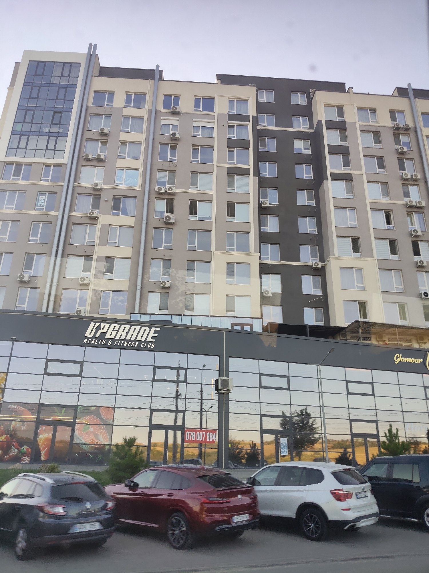 The image shows a modern, multi-story building with a glass facade on the ground floor. The building appears to be located in Chișinău, Moldova, as indicated by the text "UPGRADE HEALTH & FITNESS CLUB" and "Glamour" visible on the facade, and a phone number starting with "078" which is a Moldovan mobile prefix. Several cars are parked in front of the building, including a dark grey hatchback, a burgundy coupe, a white SUV, and a black SUV. The sky is overcast. The building's architecture is contemporary, with a mix of grey, beige, and dark grey sections. Large windows and air conditioning units are visible on the upper floors. The ground floor features large, reflective windows displaying images of food items, possibly indicating a restaurant or market. The overall scene is that of an urban commercial and residential area.