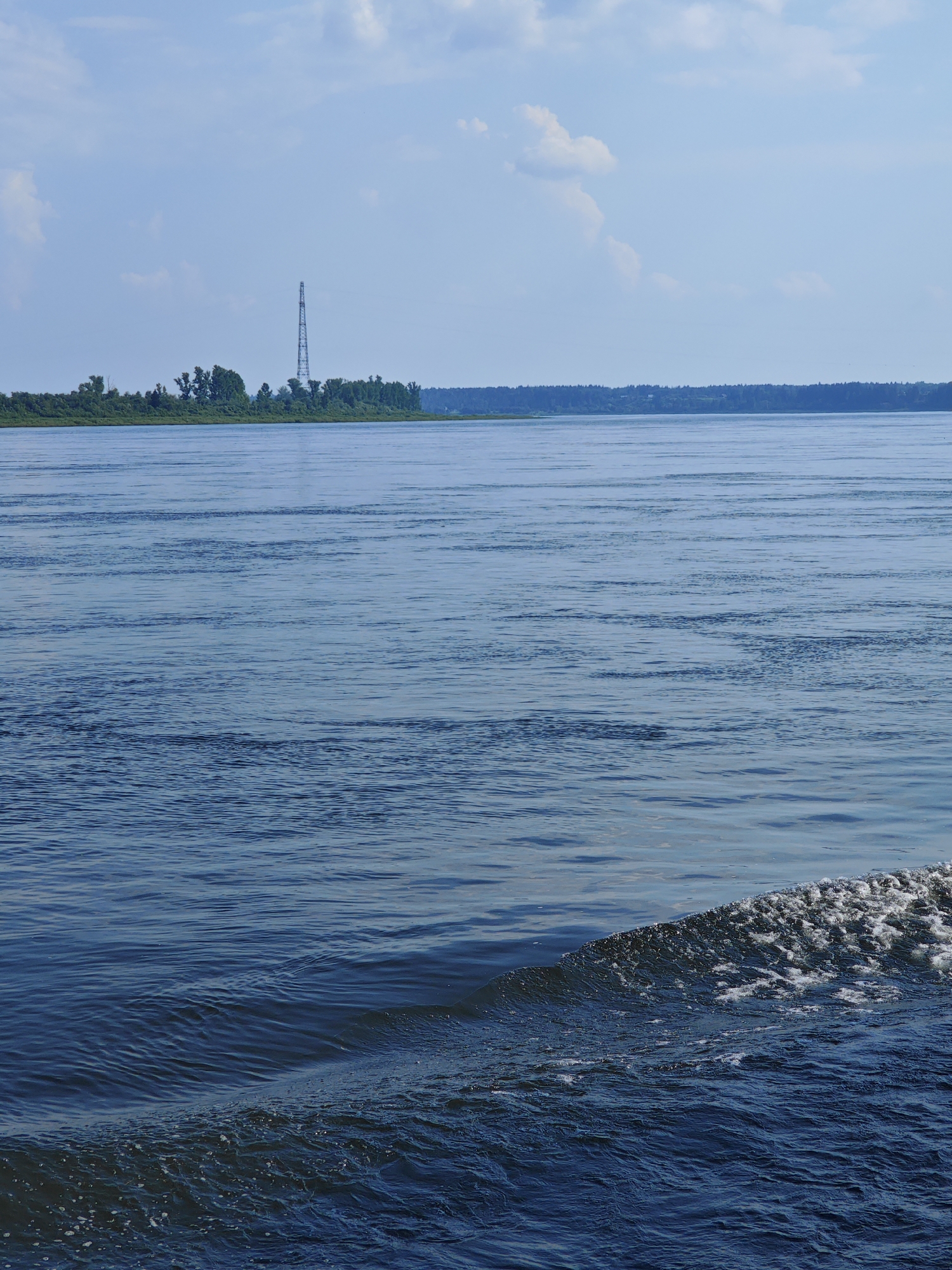 The image captures a wide, calm river on a partly cloudy day. The water is a deep blue, reflecting the sky. In the distance, a tree-lined shore stretches across the horizon, dotted with a tall, lattice-like communication tower on the left side of the frame. The foreground shows a small wave or wake breaking, creating white foam against the darker blue water. The scene appears to be a natural landscape with some man-made infrastructure in the distance.