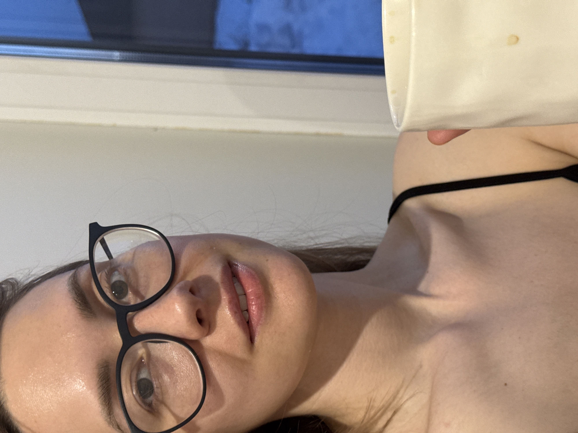 A woman with brown hair and glasses is lying down. She is wearing a black strap, likely a swimsuit top. She is holding a white mug. In the background, there is a window. There is no information that indicates that the image was taken in Chaikovsky, Russia.