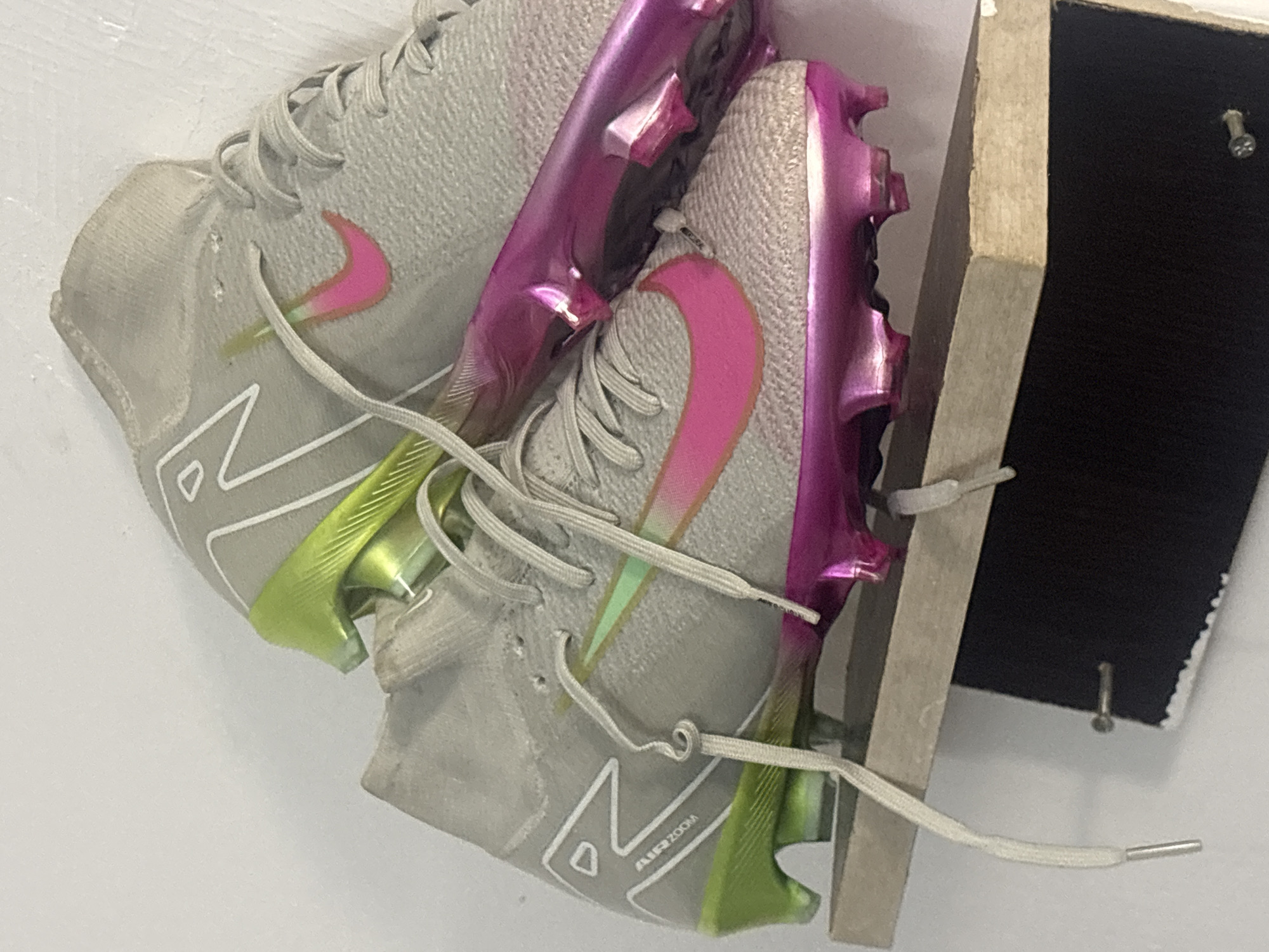 The image depicts a pair of grey and pink Nike Mercurial soccer cleats mounted on a wooden frame. The cleats are stacked on top of each other, angled towards the upper left of the frame. The wooden frame is adjacent to a dark surface. The environment appears to be indoors with white walls. There is no indication of the location being in Bukuru, Nigeria. There are no people visible in the image.