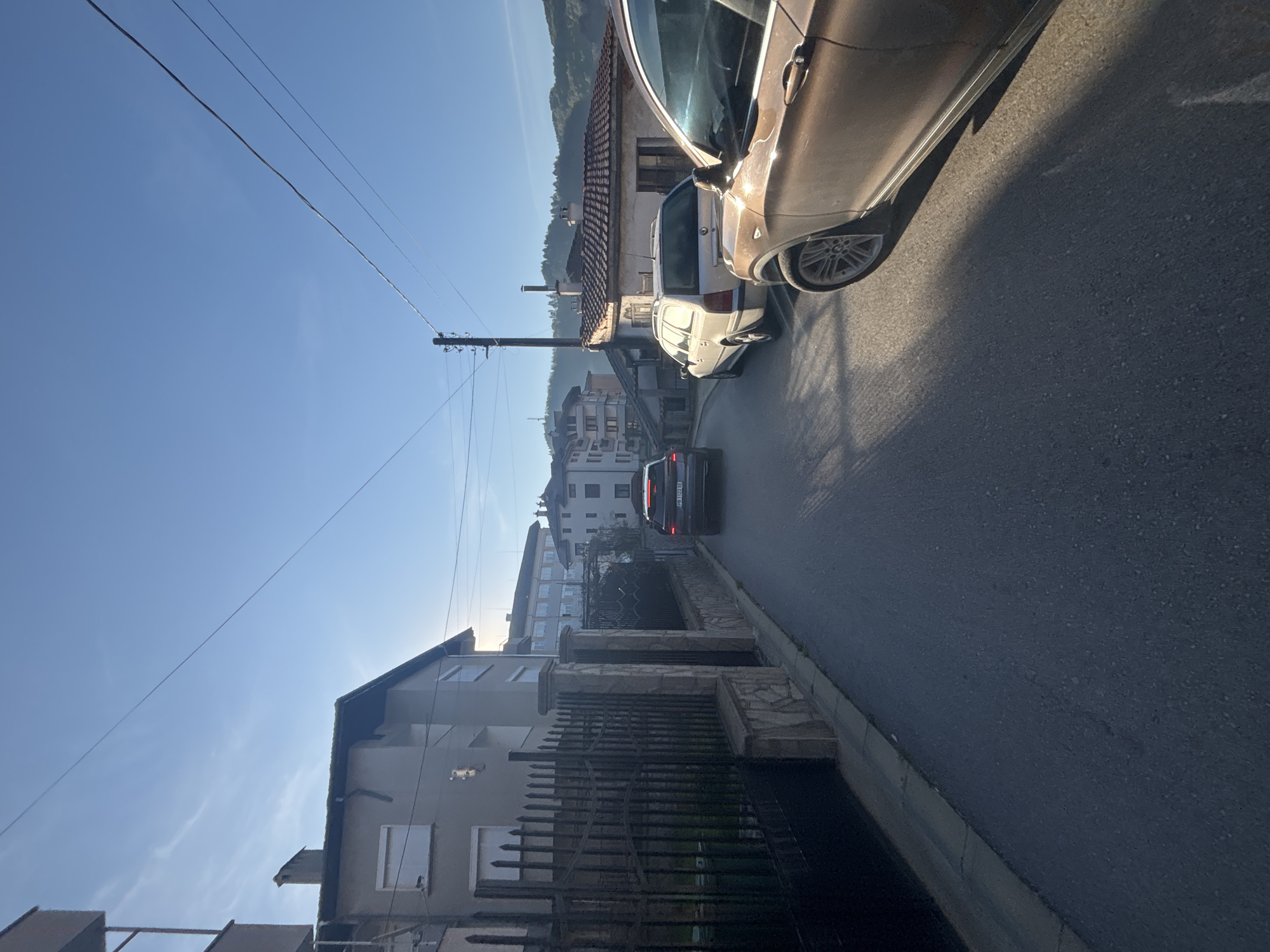 The image shows a street scene in Zlatograd, Bulgaria. The photograph is taken at street level, looking uphill. The street is lined with buildings on both sides, some with fences. A dark-colored car is driving uphill. Two parked cars, a white car and a brown car, are visible on the right side of the street. Power lines stretch across the sky. The sky is blue, and the scene appears to be illuminated by sunlight.