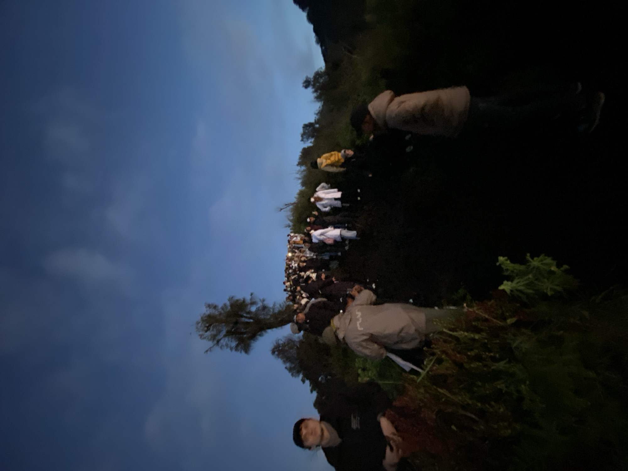 In Cemorolawang, Indonesia, a large group of people are gathered outside in a grassy area at dusk or dawn. Many are wearing white coats, some also have colored outerwear. Two people are standing closer to the foreground, while many more are visible in the distance. The sky is dark blue with some clouds. A tree is visible to the left of the group.
