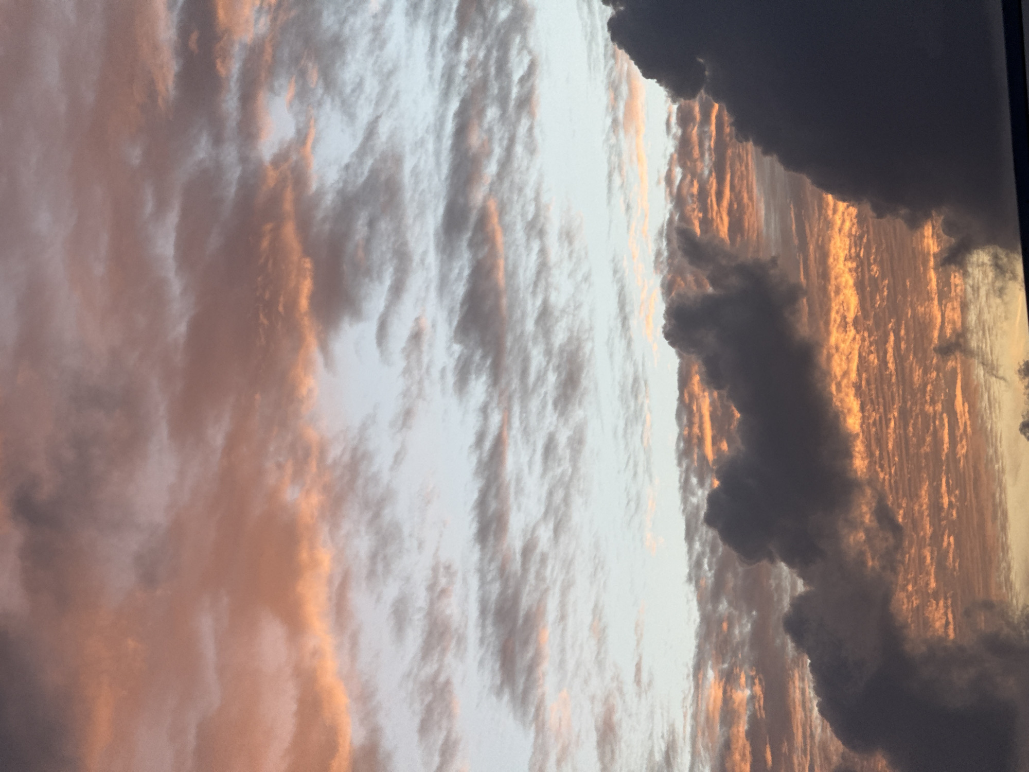 The image depicts a dramatic sunset over the ocean in Karon, Thailand. The sky is filled with a dynamic array of clouds, illuminated by the setting sun. The clouds range in color from deep, dark grays and purples in the foreground, suggesting thicker, denser formations, to lighter, wispy clouds in the background painted with vibrant oranges, pinks, and yellows. These warmer tones highlight the textured nature of the clouds, creating a rippling or layered effect. A sliver of the ocean is visible at the very bottom, appearing dark and somewhat textured. The scene is devoid of people or man-made objects, focusing solely on the atmospheric conditions and the natural beauty of the sky at dusk.