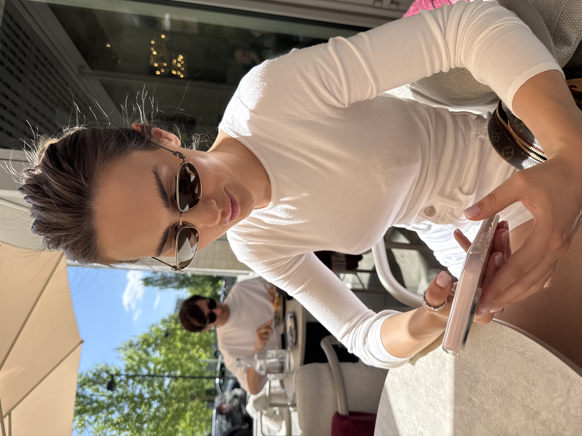 A woman with brown hair pulled back, wearing sunglasses and a white long-sleeved shirt, is seated at a table outside. She is holding a cell phone in her hands. A reflection of another person wearing sunglasses and a white shirt can be seen in the background. The scene appears to be an outdoor café or restaurant with an umbrella providing shade.