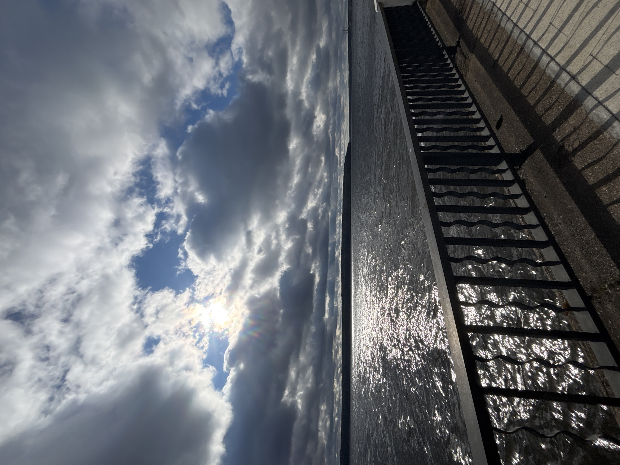 The image captures a panoramic view of a large body of water alongside an embankment, under a dynamic sky. The sky, occupying the left half of the frame, features a mix of bright white and dark grey clouds, with patches of clear blue sky visible. The sun is partially obscured by clouds in the mid-left, casting intense rays and a slight lens flare, indicating its low position.
The center of the image is dominated by the dark, expansive water, likely a river or reservoir. The surface of the water intensely reflects the sunlight, creating shimmering, silver patterns, particularly noticeable towards the right.
On the right side of the frame, a concrete embankment or wall runs horizontally, separating the water from the land. A dark metal structure, resembling a wide ladder or a set of steps with wavy rungs, extends from the embankment towards the water's edge. Strong, elongated shadows from this structure and the embankment are cast onto a lighter, textured ground surface adjacent to the water, suggesting either early morning or late afternoon. A faint, dark line on the distant horizon separates the water from the sky, hinting at a far shoreline. The scene portrays a waterfront area in Chaikovsky, Russia, likely the Votkinsk Reservoir. No specific activity or text is visible.