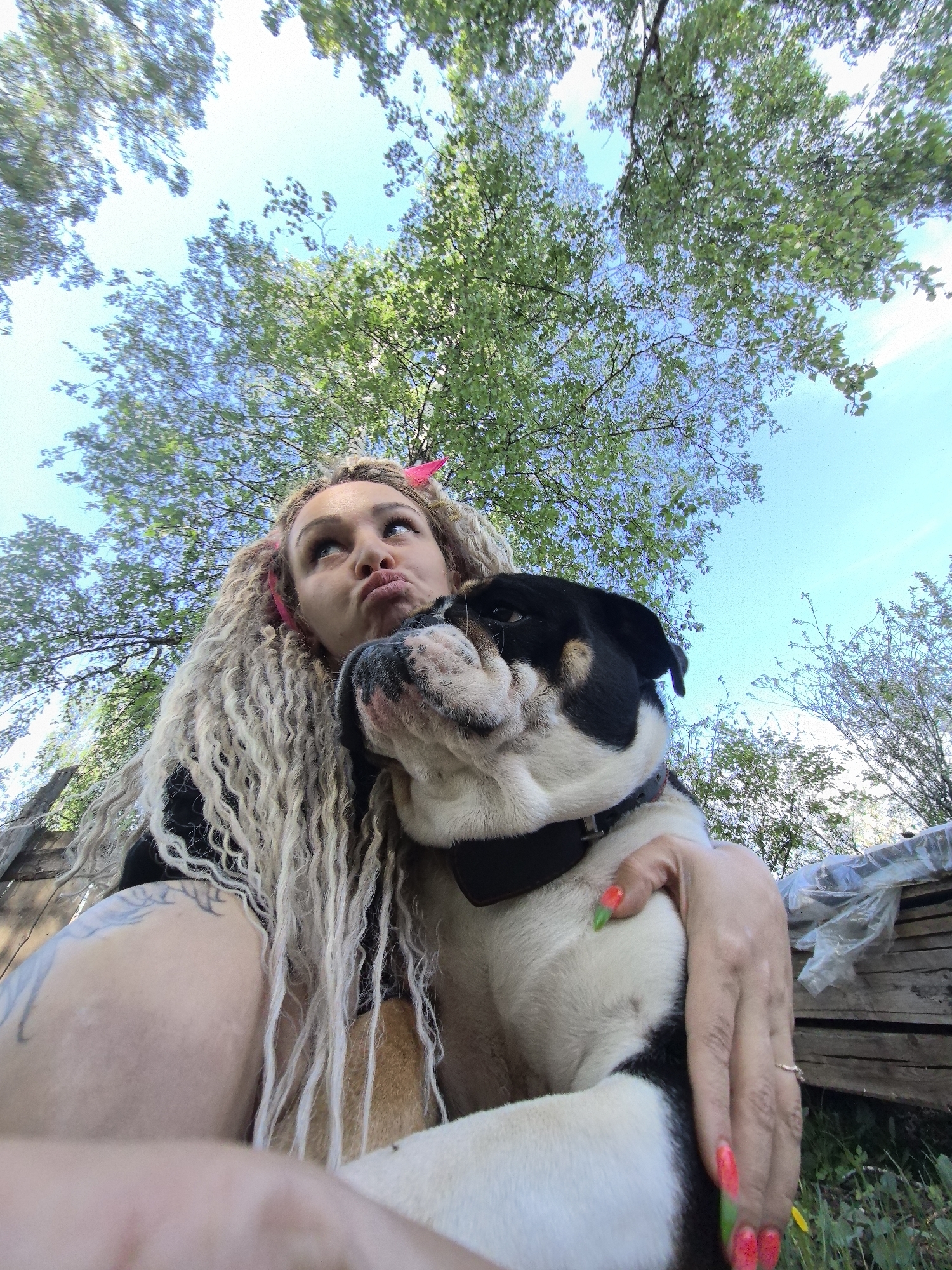 A woman and a dog are the primary subjects of this outdoor, low-angle photograph. The woman, with long blonde curly hair secured with pink hair ties, is making a pouting facial expression and looking upwards. She is wearing a black top and has tattoos on her arm and upper chest. Her fingernails are painted with a red and green design. The dog, a black and white bulldog with a black collar, is nestled in the woman's arms.
The background features a partially obscured view of trees with green leaves and a light blue sky with some clouds. In the lower left, a wooden structure is visible. The location is described as Noviy, Russia. The action is an embrace between the woman and the dog.