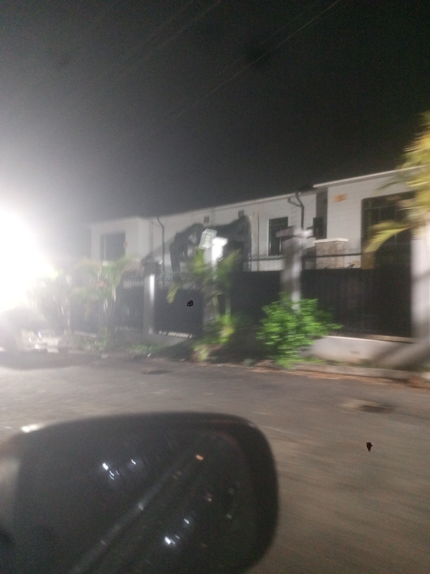 The image depicts a residential street scene at night in Gwarinpa, Nigeria. A white, single-story building with dark windows and a dark metal gate is visible behind foliage. The scene is partially obscured by bright lights on the left and blurriness throughout the image. The side mirror of a vehicle is in the foreground, and the road surface is visible, with a dark object on it. The sky is dark, with visible power lines.