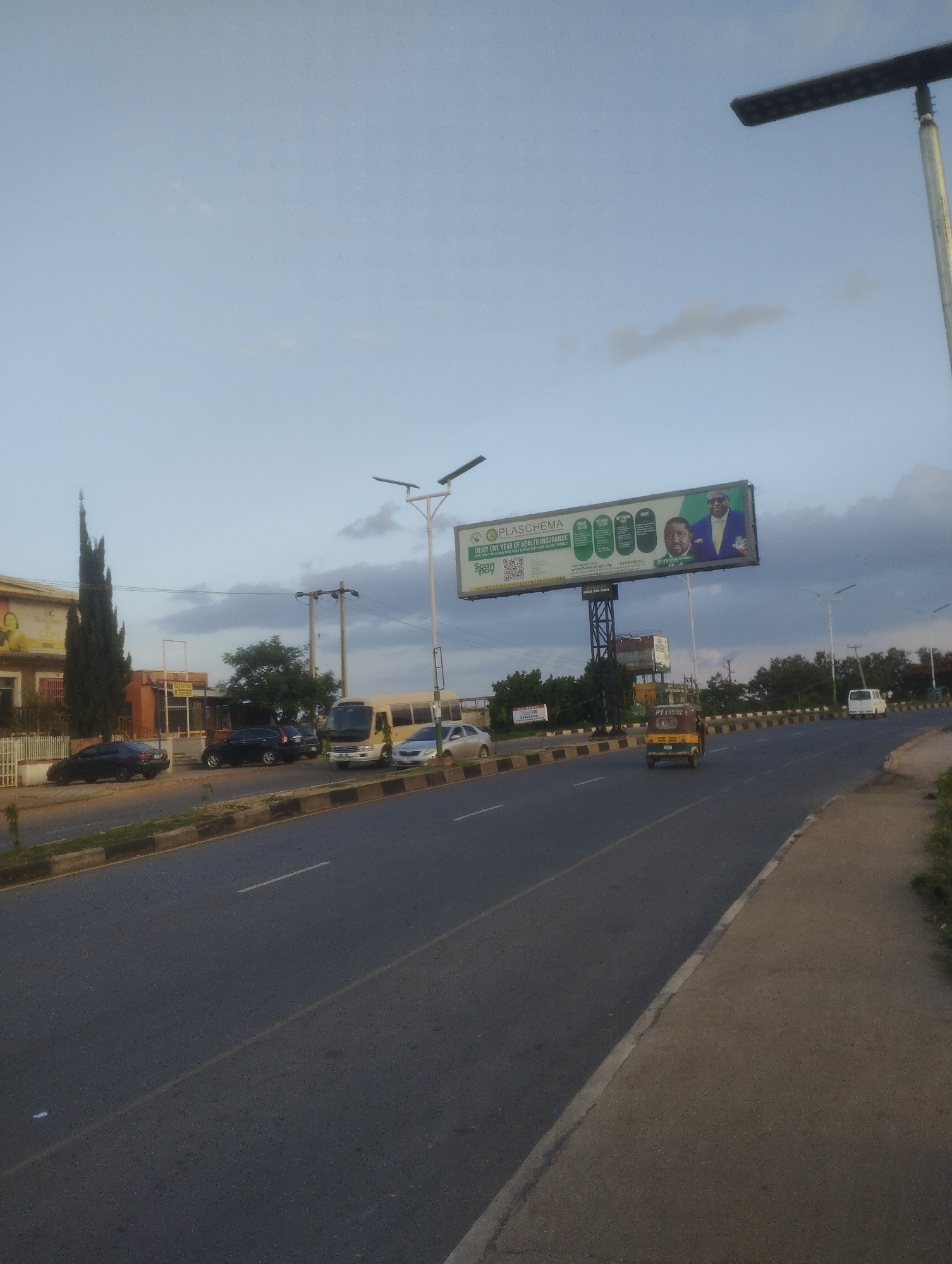 The image captures a street scene in Jos, Nigeria, at what appears to be either dawn or dusk, given the overcast sky and soft lighting. The foreground shows a paved road with distinct lane markings, including a solid white line and a broken yellow line. A concrete sidewalk with a curb runs along the right side of the road.
In the mid-ground, several vehicles are visible: a dark sedan and a black SUV parked on the left side of the road, a white bus, and a silver sedan driving on the road. Further down the road, a yellow and green three-wheeled auto-rickshaw is moving away from the camera. A large billboard advertising "PLASCHEMA" and "ENJOY ONE YEAR OF HEALTH INSURANCE" is prominently displayed in the background, featuring images of two men and QR codes. To the left of the billboard, a tall, slender building with a beige facade is visible, with a partial view of a billboard featuring a woman.
The environment is characterized by a mix of urban infrastructure and vegetation. Streetlights with solar panels are mounted on poles along the road. Trees, including a very tall, narrow cypress-like tree, are interspersed with buildings and the road. The sky is cloudy, with shades of blue and grey. The overall impression is of a typical urban street in Jos, Nigeria, with active traffic and commercial advertising.