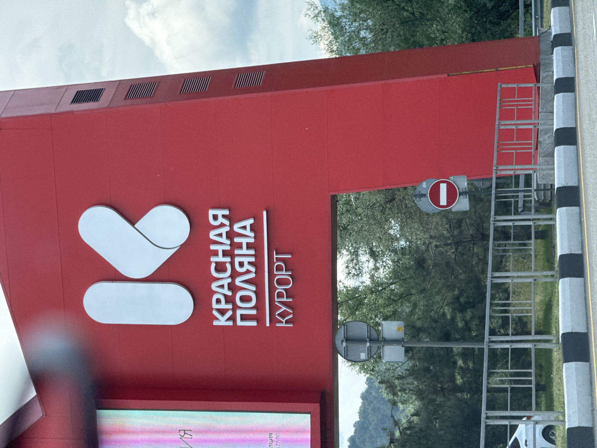 The image shows a large red archway in Krasnaya Polyana, Russia, with the Cyrillic text "КРАСНАЯ ПОЛЯНА КУРОРТ" (Krasnaya Polyana Resort) in white letters. A stylized white "K" logo is prominently displayed on the arch. To the left of the arch is a large digital billboard with colorful, blurred content. The archway is situated over a road with a black and white striped curb. A metal fence separates the road from a grassy area with trees and mountains in the background. A traffic sign with a red circle and a white horizontal bar, indicating "no entry," is visible on a pole near the arch. The sky is overcast with some clouds. There are no people visible in the image.