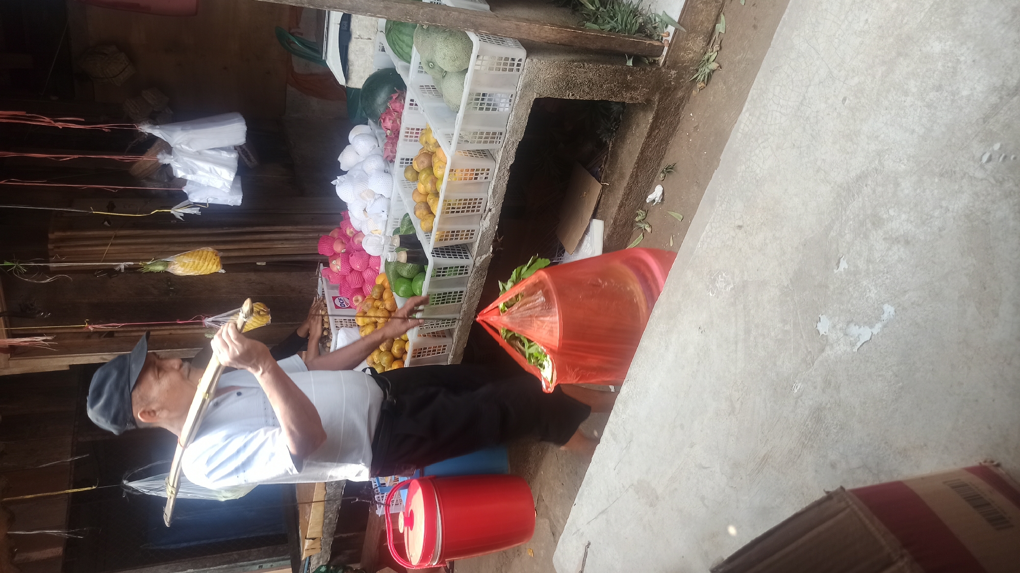 An elderly Asian man wearing a black cap and a white short-sleeved shirt is walking in a market stall in Tuba, Indonesia. He is carrying a long pole over his shoulders, with two pineapples hanging from it. The man is walking from left to right and appears to be facing away from the camera. In the background, there is a display of fruits, including oranges, avocados, and watermelons. There are also plastic bags hanging from the ceiling. To the man's left, there is a large red bucket. In front of the man, there is a large orange plastic bag filled with green vegetables. The ground is made of concrete, and there is some debris on the ground. The scene appears to be outdoors, with natural light coming from the top. The man's attire and the market setting suggest he is a vendor or a customer.