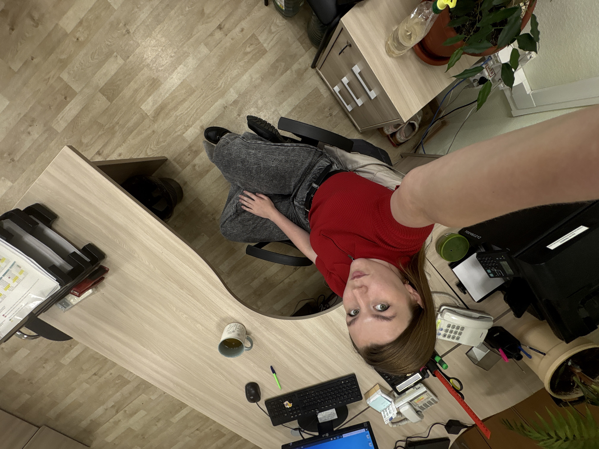 A medium-shot, high-angle photograph depicts a person in an office setting. The person is wearing a red, long-sleeved shirt and grey jeans, seated on a black office chair. The office space contains a light-colored wooden desk with a computer monitor, keyboard, mouse, a pen, a mug, a telephone, printer, and various office supplies. Also visible is a potted plant, a water bottle, and a small cabinet. The floor is covered with light-colored wooden planks. No information about Izhevsk, Russia can be confirmed from the image.