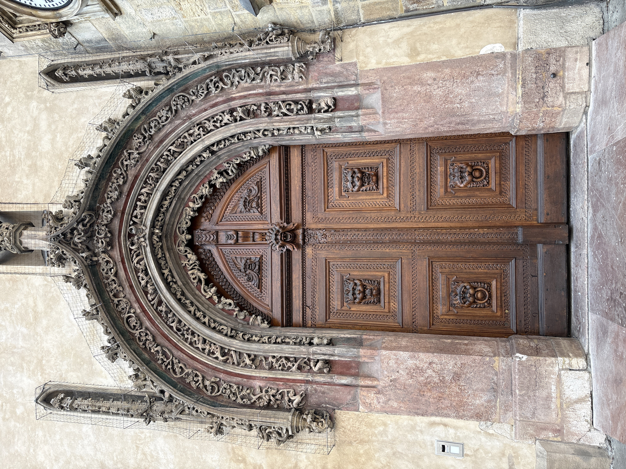 The image shows an ornate wooden double door set within a highly decorated gothic archway. The archway is carved with intricate details, including foliage, figures, and gargoyle-like faces. The doorway is flanked by stone pillars. The door itself is made of dark wood and features carved panels, some with lion head motifs and ring handles. Above the door, a carved face with an open mouth is visible. The facade surrounding the archway is a light-colored plaster. A wire mesh is visible attached to some of the architectural elements, suggesting ongoing restoration or protection work. The ground in front of the door is paved with stone tiles. A clock face is partially visible on the right side of the building. This is the entrance to the Old Town Hall Tower in Prague, Czechia.