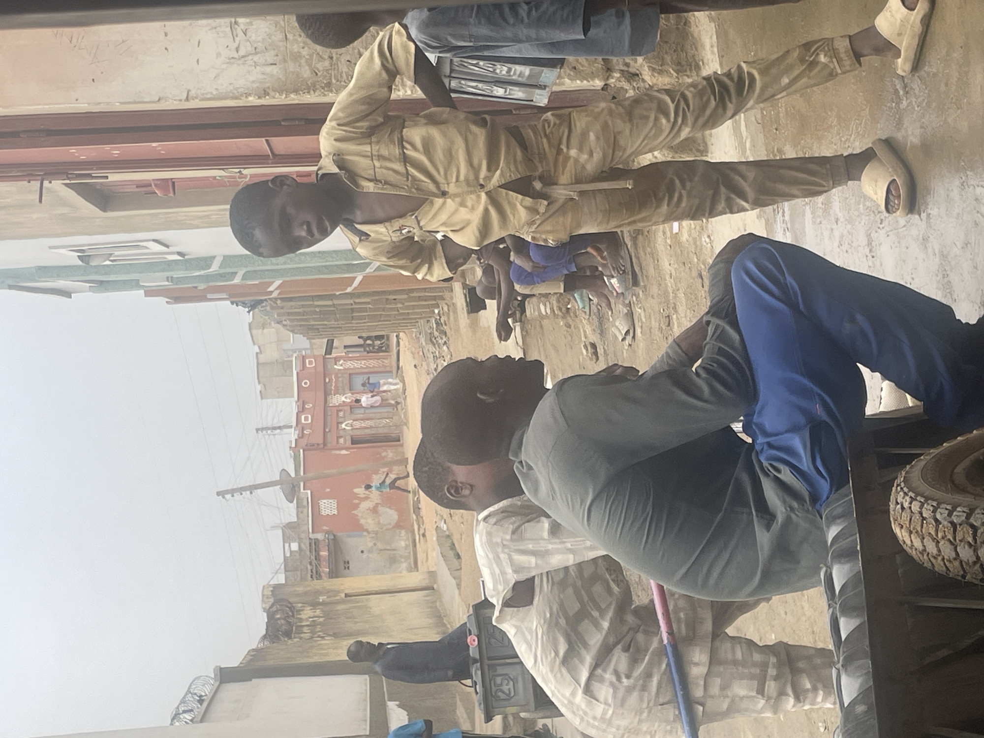 Several individuals are visible in this outdoor scene. One individual, dressed in a tan jumpsuit and sandals, lies prone on the ground. Another person, wearing a grey shirt and blue pants, is seated in what appears to be a vehicle. Several other people are situated in the background. The setting seems to be a street in an urban area, with buildings visible in the backdrop, and utility poles and wires overhead.