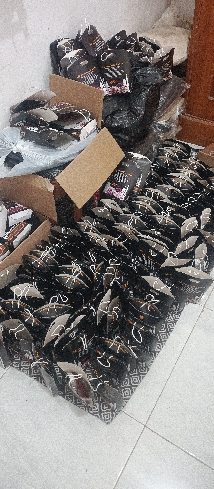 In Arjasa, Indonesia, a collection of dark brown gift bags with white rope handles is neatly arranged on a patterned rug. The bags are adorned with Islamic calligraphy and graphics of mosques and dates, indicating their purpose as souvenirs from Hajj or Umrah pilgrimages. Some bags are open, revealing small pouches containing what appear to be prayer beads or dates. The bags are stacked in rows, creating a dense display. In the background, several cardboard boxes and large black plastic bags are visible, suggesting a storage or preparation area. The scene appears to be indoors, with a plain white wall and a wooden doorframe in the upper right corner. The lighting is diffuse, casting soft shadows on the floor.