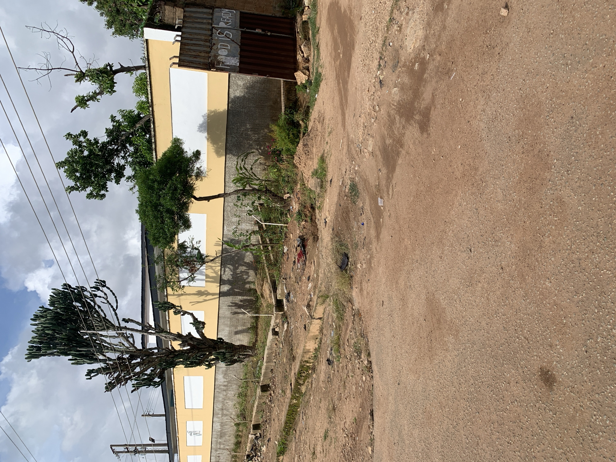 A wide shot captures a dusty, uneven dirt road leading towards a long, yellow-and-white striped wall topped with barbed wire. The wall is flanked by a large, multi-branched cactus on the left and several smaller trees and bushes. A rusty metal gate with the words "FOOD IS READY" painted on it stands open on the right side of the wall. The ground is littered with debris, including plastic bags, rocks, and what appears to be a pile of red fabric. The sky is overcast with clouds.