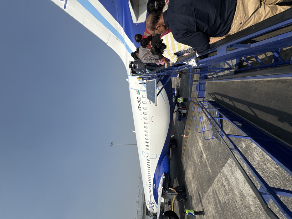 A line of people ascend an aircraft boarding bridge, walking towards the open door of a blue and white airplane. The plane is marked with the registration "VT-IRZ" and features a solar panel near the front. An airport worker in a neon vest is visible on the tarmac beside the plane. The sky is clear and blue. The perspective is from ground level looking upward.