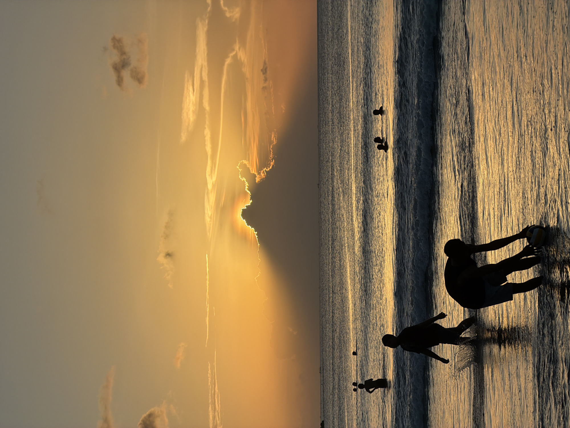 This image captures a scene at Kuta Selatan, Indonesia, during a sunset. The sky exhibits a gradient of colors, from a pale blue to a deep orange where the sun is obscured behind clouds, casting crepuscular rays. The ocean stretches towards the horizon, reflecting the colors of the sky. Silhouetted figures of people populate the shoreline. Two individuals are interacting, one holding a volleyball, suggesting a game. Further out, several smaller silhouettes are visible, likely representing people standing in the water. The water itself is textured with small waves catching the light.