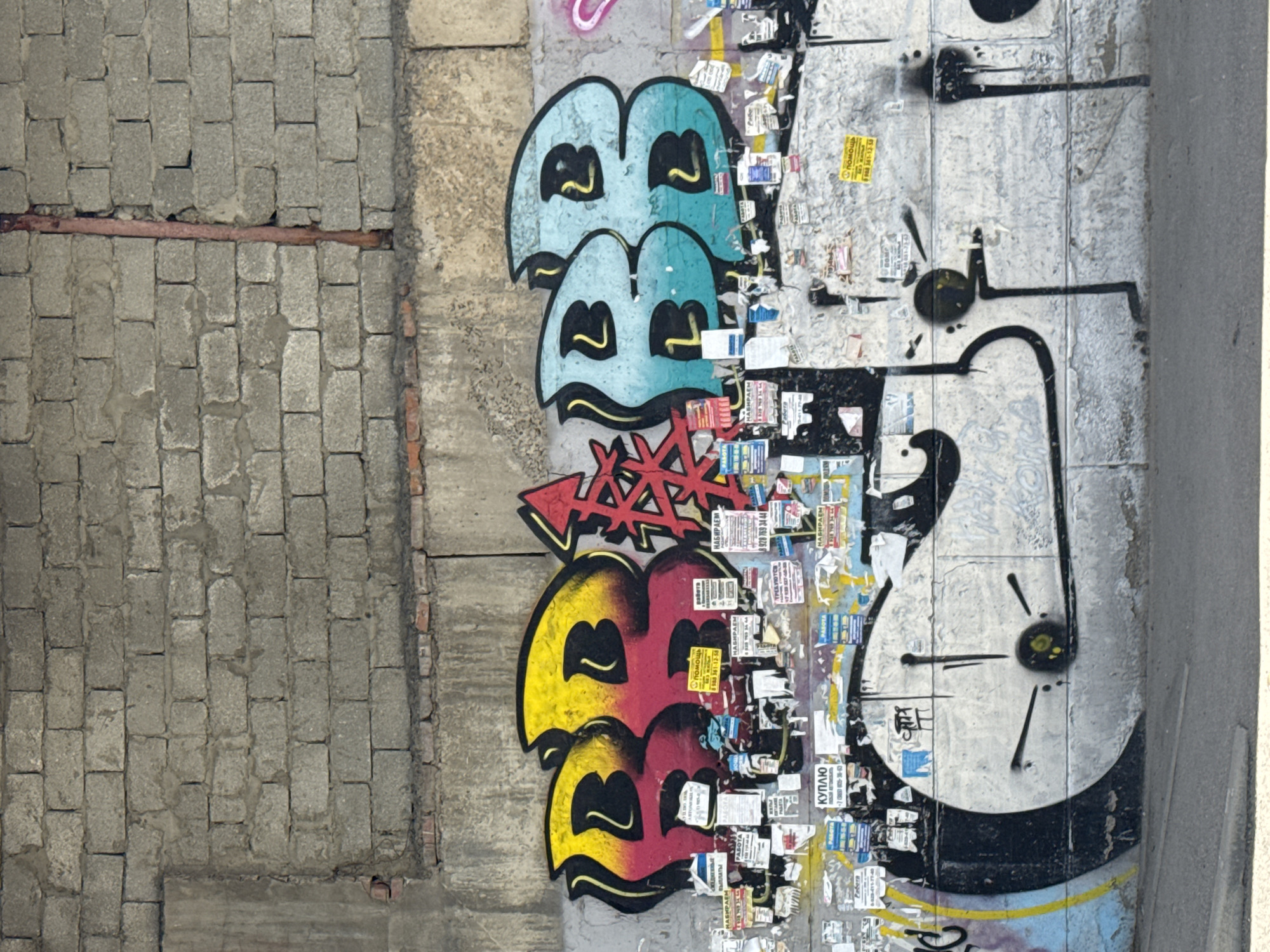 The image shows a vertical view of a concrete wall in what appears to be an urban environment, heavily covered in graffiti and posters.
On the left side of the image is a section of the wall made of grey bricks.
The right side of the image is covered in layers of graffiti and overlaid with numerous posters. The graffiti includes a series of cartoonish, stacked blue and yellow/red "B" shapes, some of which have a distressed aesthetic. There is also a white graffiti design that seems to resemble a cartoonish clock face with short, thick black lines emanating from a central point.
Numerous posters are pasted over the graffiti, particularly between and around the "B" shapes and the clock graffiti. The posters appear to be Russian as a few words from the OCR text are in Russian and Cyrillic characters are visible.
Based on the OCR text and the visible posters, this scene is likely in the city of Krasnodar, Russia.