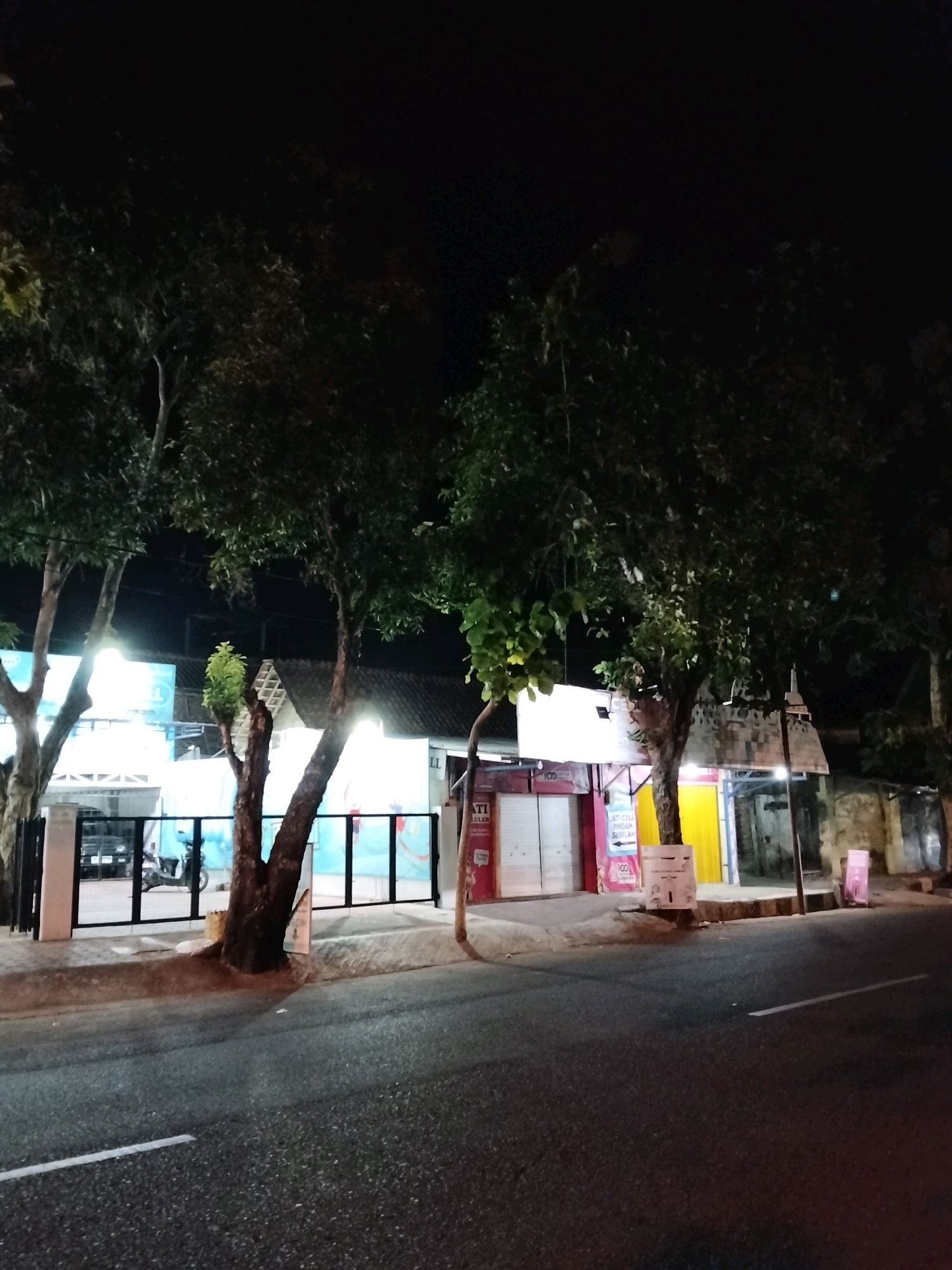 The image depicts a nighttime scene on a paved road in Purworejo, Indonesia. The road is bordered by a sidewalk and several storefronts, suggesting a commercial area. Trees with dense foliage line the sidewalk, their branches reaching towards the dark night sky. The storefronts are illuminated by artificial lights, casting a glow on the facades and the surrounding area. Some of the storefronts have signage, though the text is not clearly legible. Parked vehicles, including a motorcycle, are visible behind a fence in front of one of the buildings. The overall impression is of a quiet, illuminated street in a city at night.