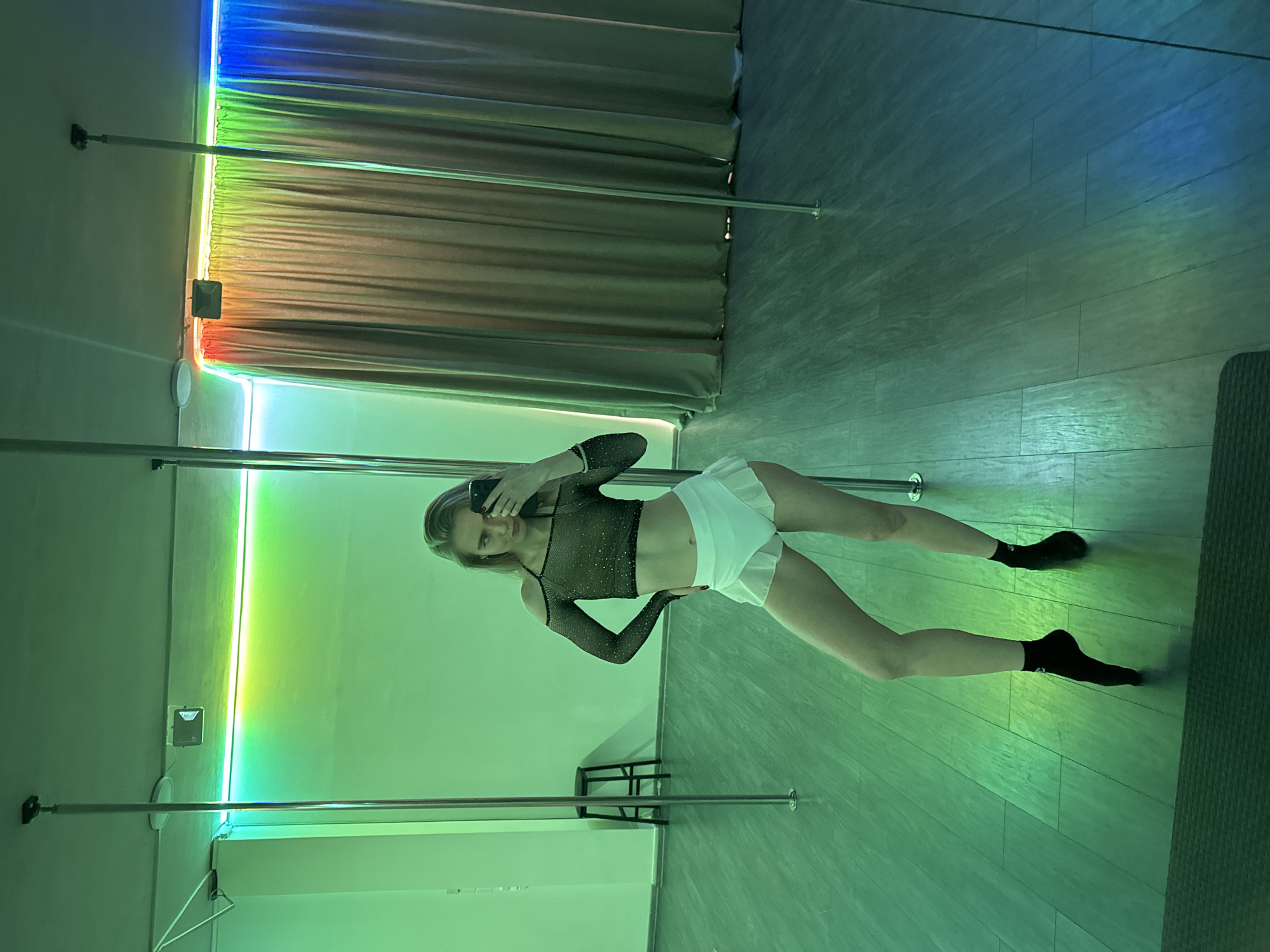 A woman is standing between two silver poles in an indoor setting. She is wearing a black, long-sleeved mesh top, a white skirt, and black socks. She is holding a cell phone up to her face, presumably taking a picture. Behind the poles is a wall and a curtain lit with colored lights. A small black stool is between the poles. The floor is plank-like and reflects the colored light.