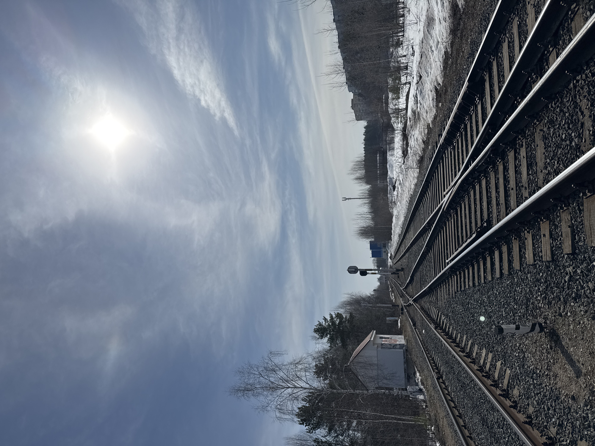 The image depicts a railway line extending into the distance under a bright, slightly overcast sky. Sunlight is prominent in the upper left. The tracks are lined with gravel ballast and run adjacent to a snowy landscape with bare trees and vegetation. There's a small building or structure to the left of the tracks, closer to the foreground. Further down the track, a railway signal and other infrastructure elements are visible. The scene appears to be taken during winter or early spring due to the presence of snow. The location is near the city of Chaikovsky, Russia based on geotagging of the image.
